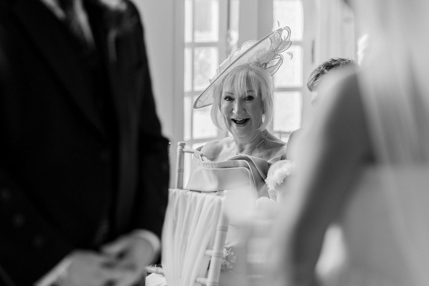 Black and white photo of a smiling woman in a fascinator hat at a wedding ceremony.