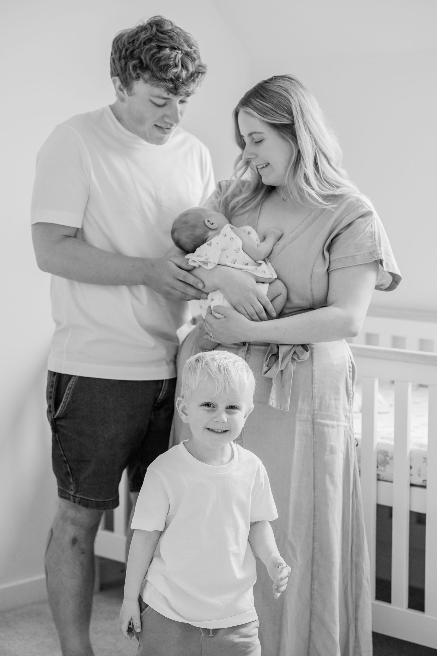 Black and white family photo of parents with newborn baby and young toddler son standing in nursery room.