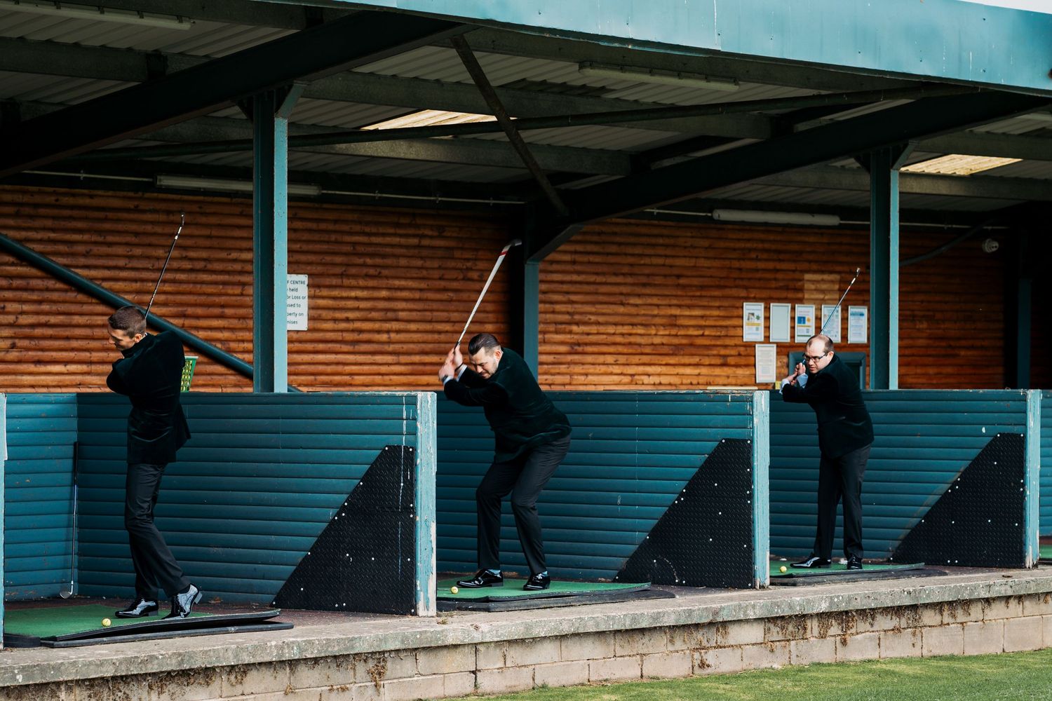 Three golfers practicing their swing at a covered driving range with teal barriers and brick backdrop.