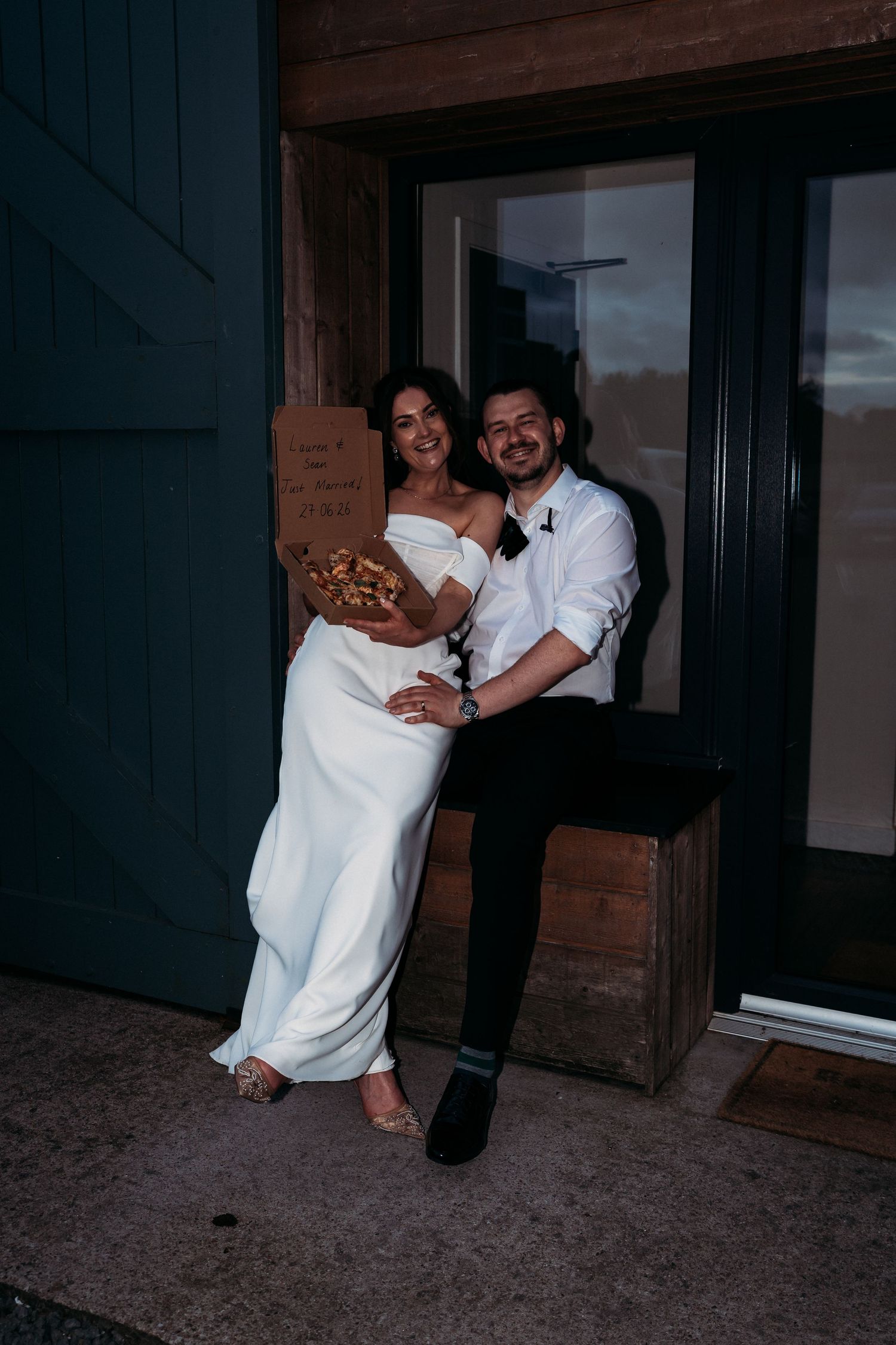 Bride in white gown and groom in white shirt share pizza outside barn venue at night during wedding reception.