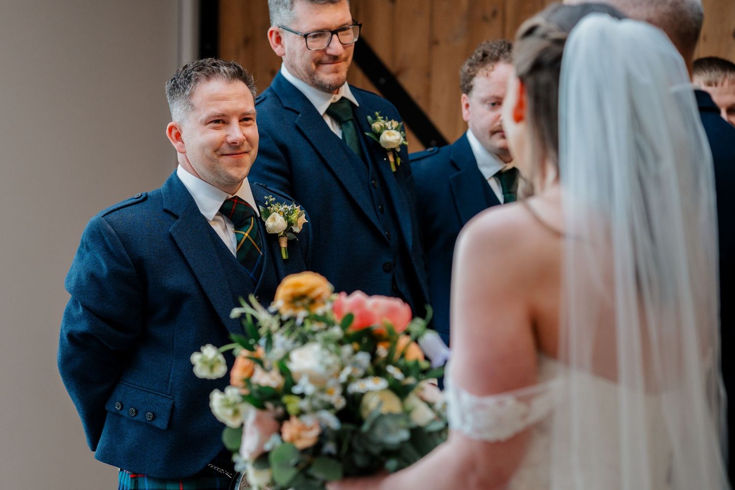Groom smiling at bride during wedding ceremony, groomsmen in navy suits standing behind, bride holding colorful bouquet.