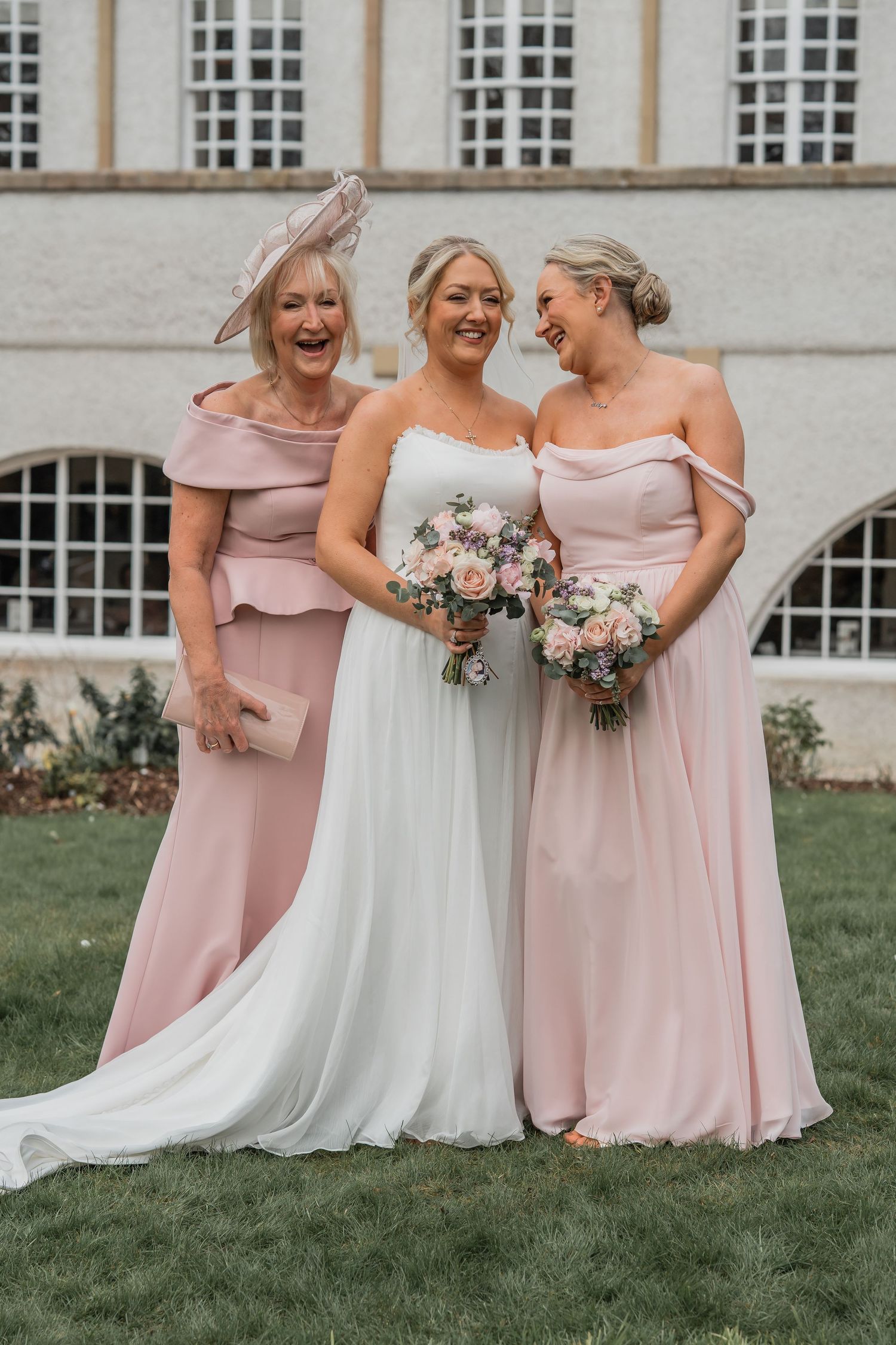 Bride in white gown with two bridesmaids in blush pink off-shoulder dresses, laughing outdoors at a wedding venue.