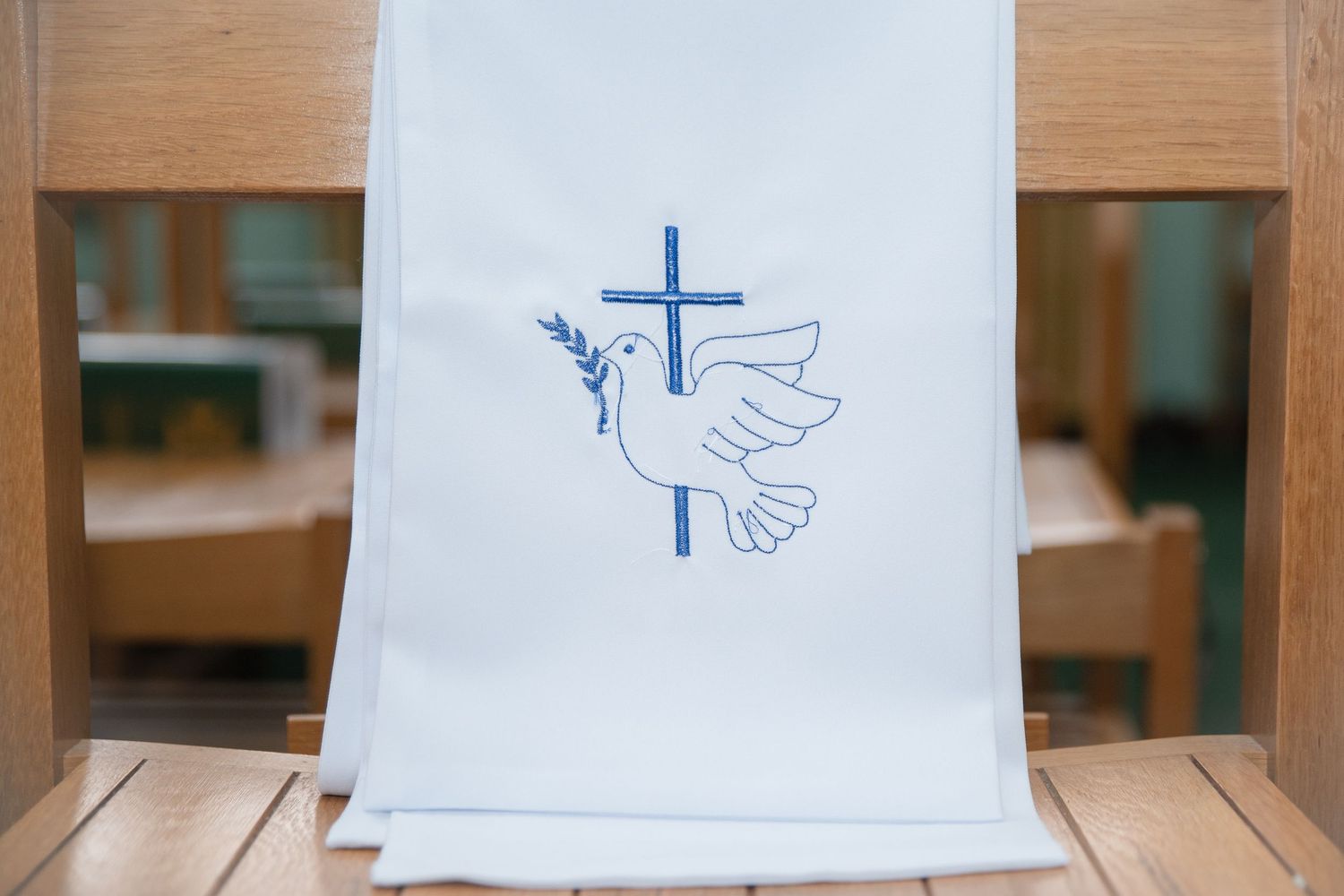 White baptism cloth with blue embroidered dove and cross symbol draped over wooden church pew.