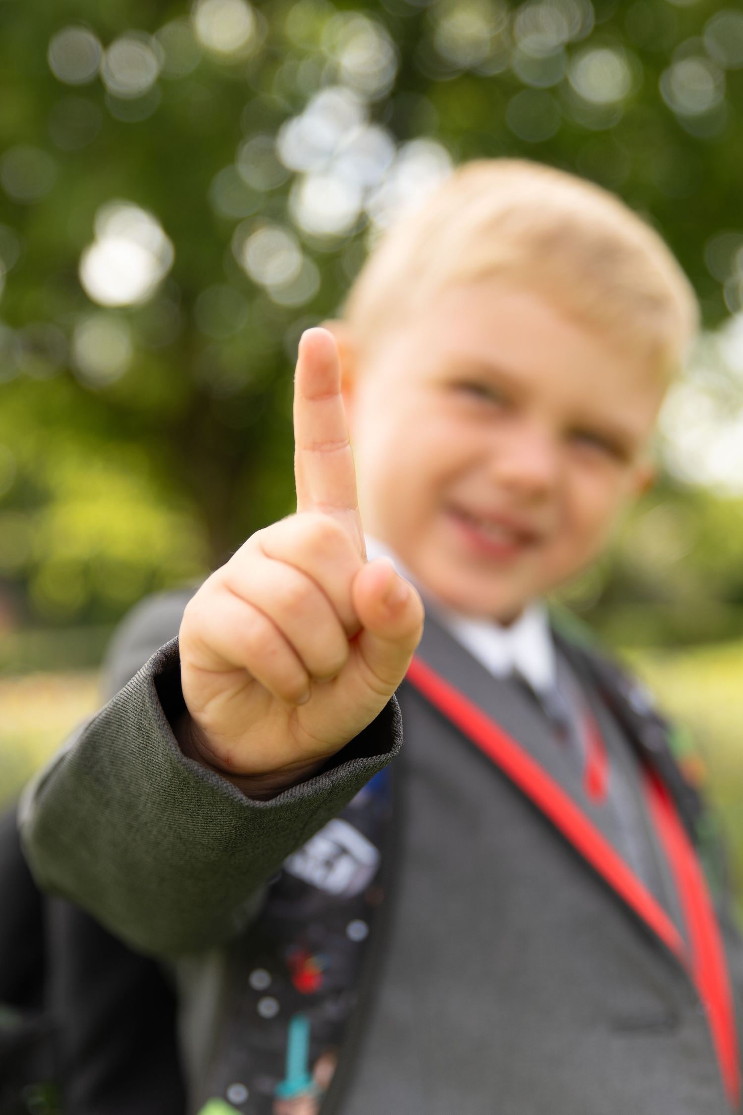 A smiling blond boy in school uniform holds up one finger outdoors with green trees in the background.