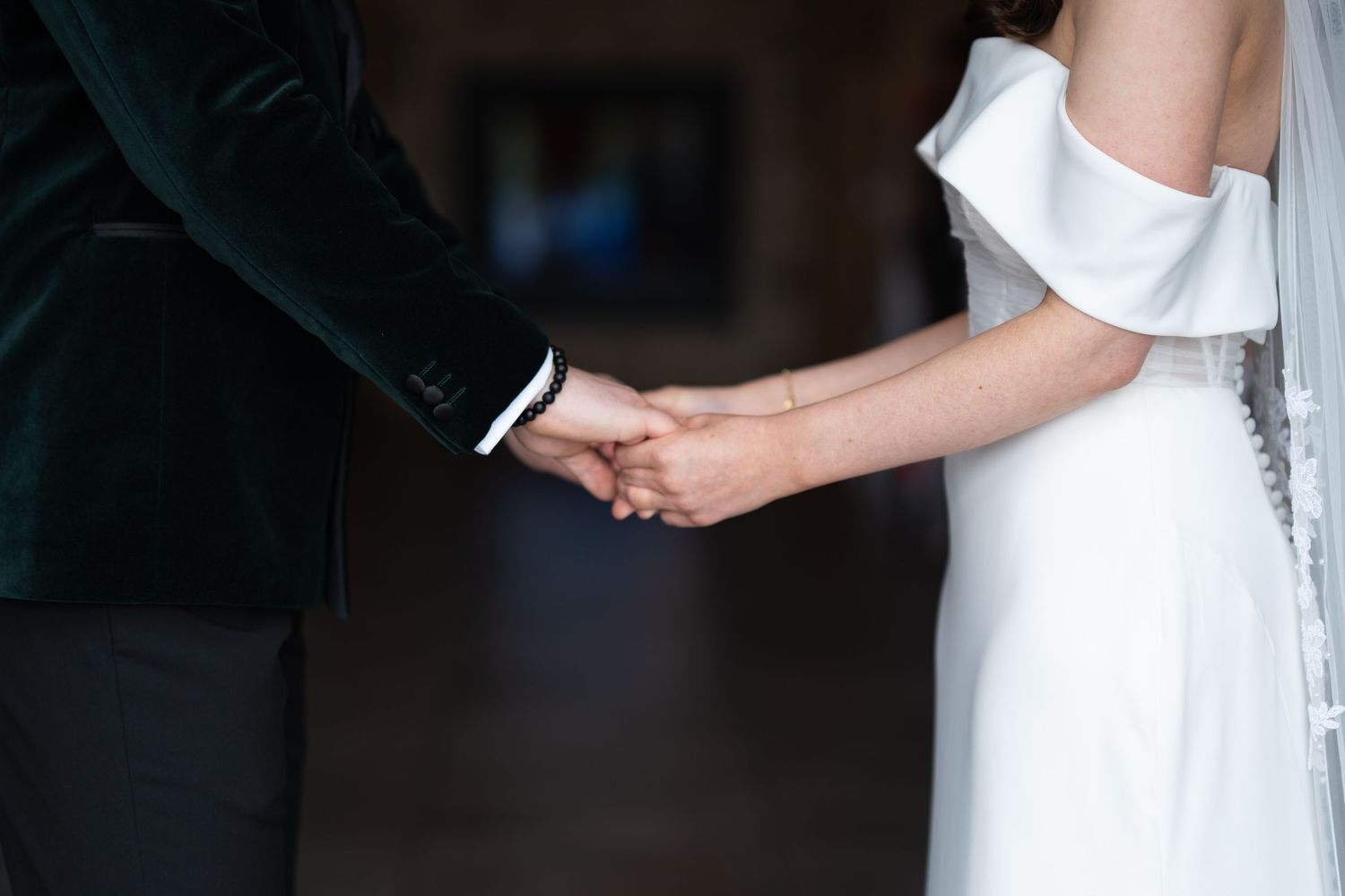 Bride and groom holding hands at wedding ceremony, bride in white off-shoulder gown, groom in dark green suit.
