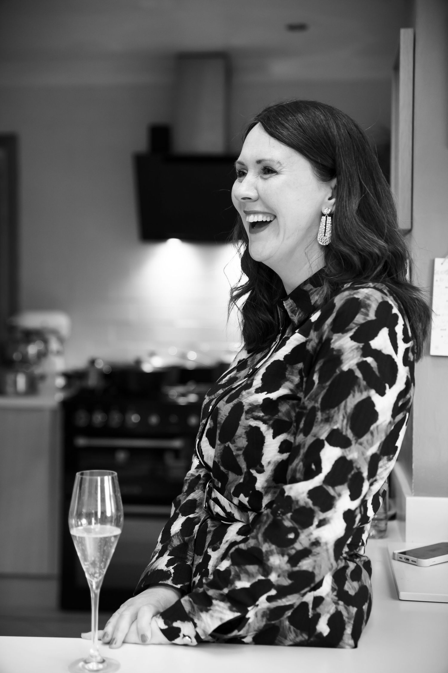 Black and white photo of a laughing woman in patterned dress holding champagne glass in a modern kitchen.