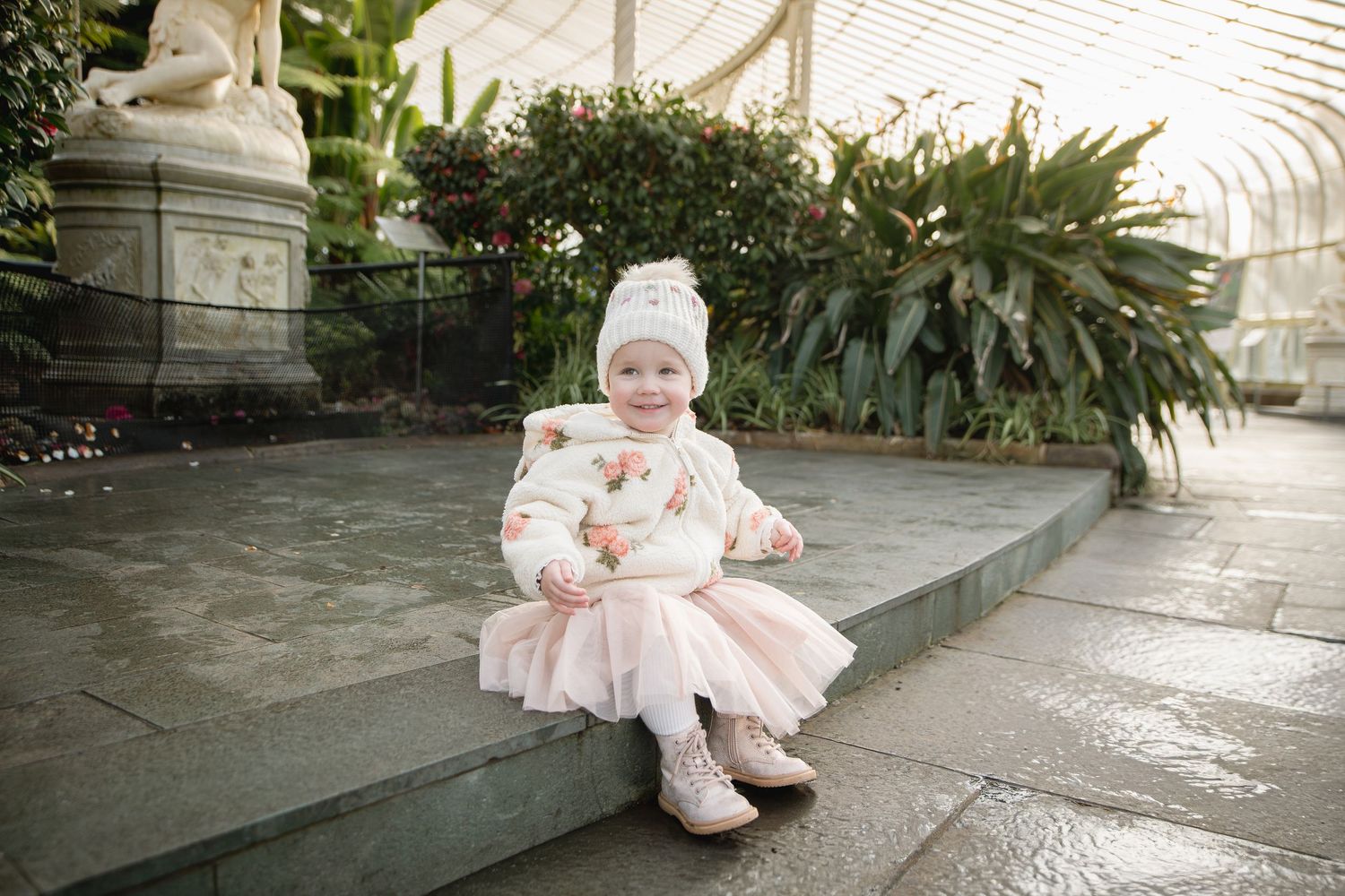Adorable toddler girl in pink tutu and white knit hat sitting on stone steps in a lush greenhouse garden.
