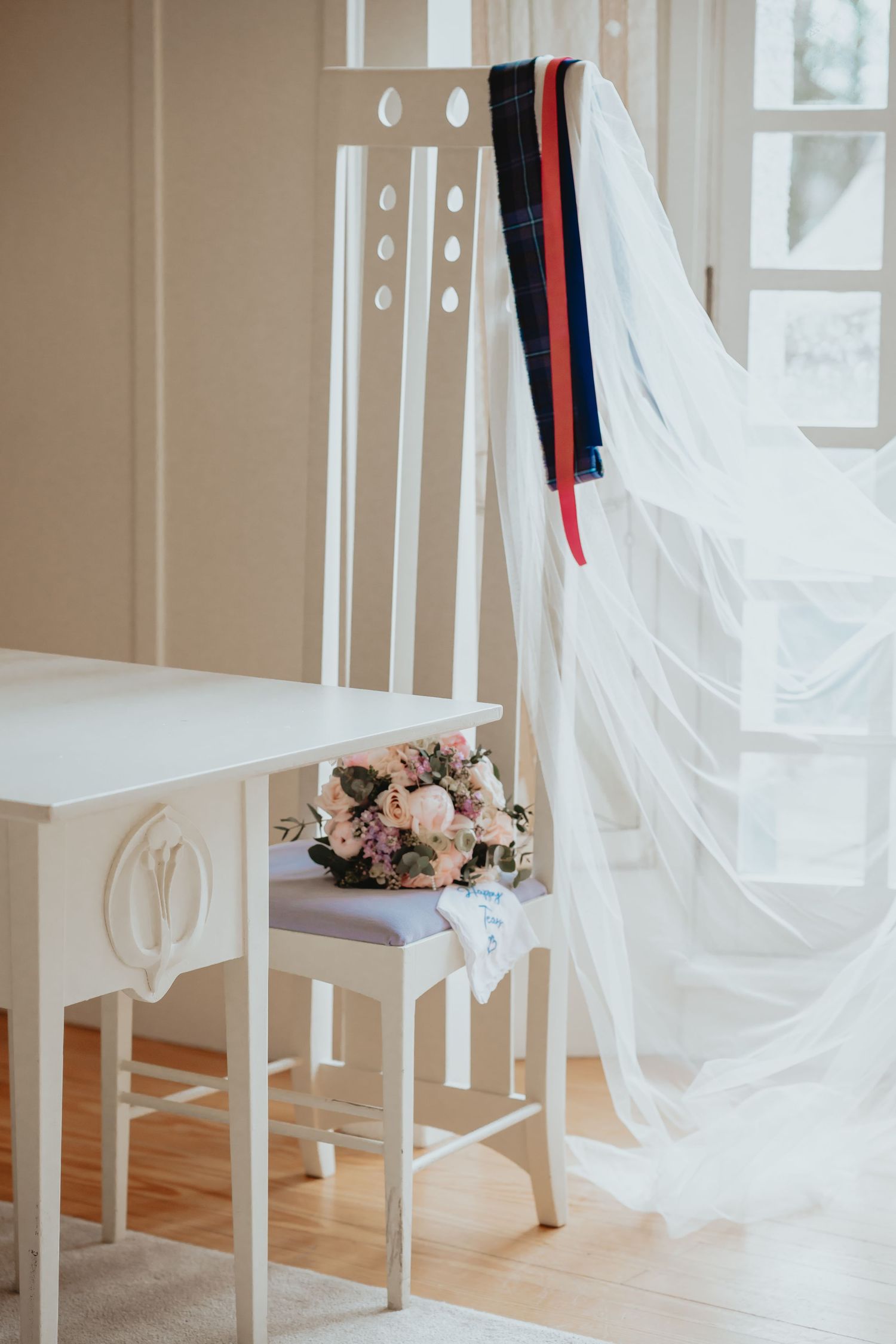 Elegant white wedding chair with bridal veil, floral bouquet, and colorful ribbons in a bright room.