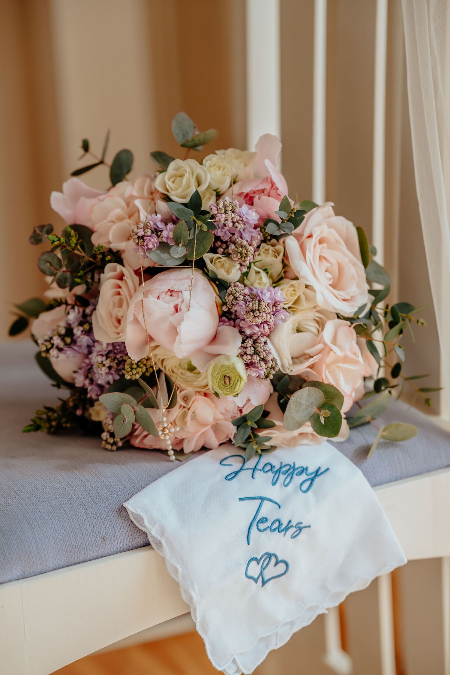 Bridal bouquet of pink peonies and roses beside an embroidered Happy Tears handkerchief on a white chair.
