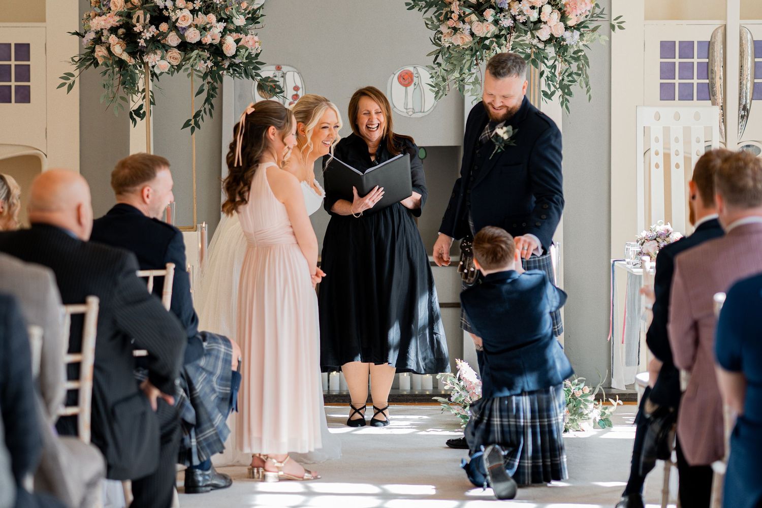 Elegant wedding ceremony with bride, groom, and officiant at floral altar, guests seated in white chairs.