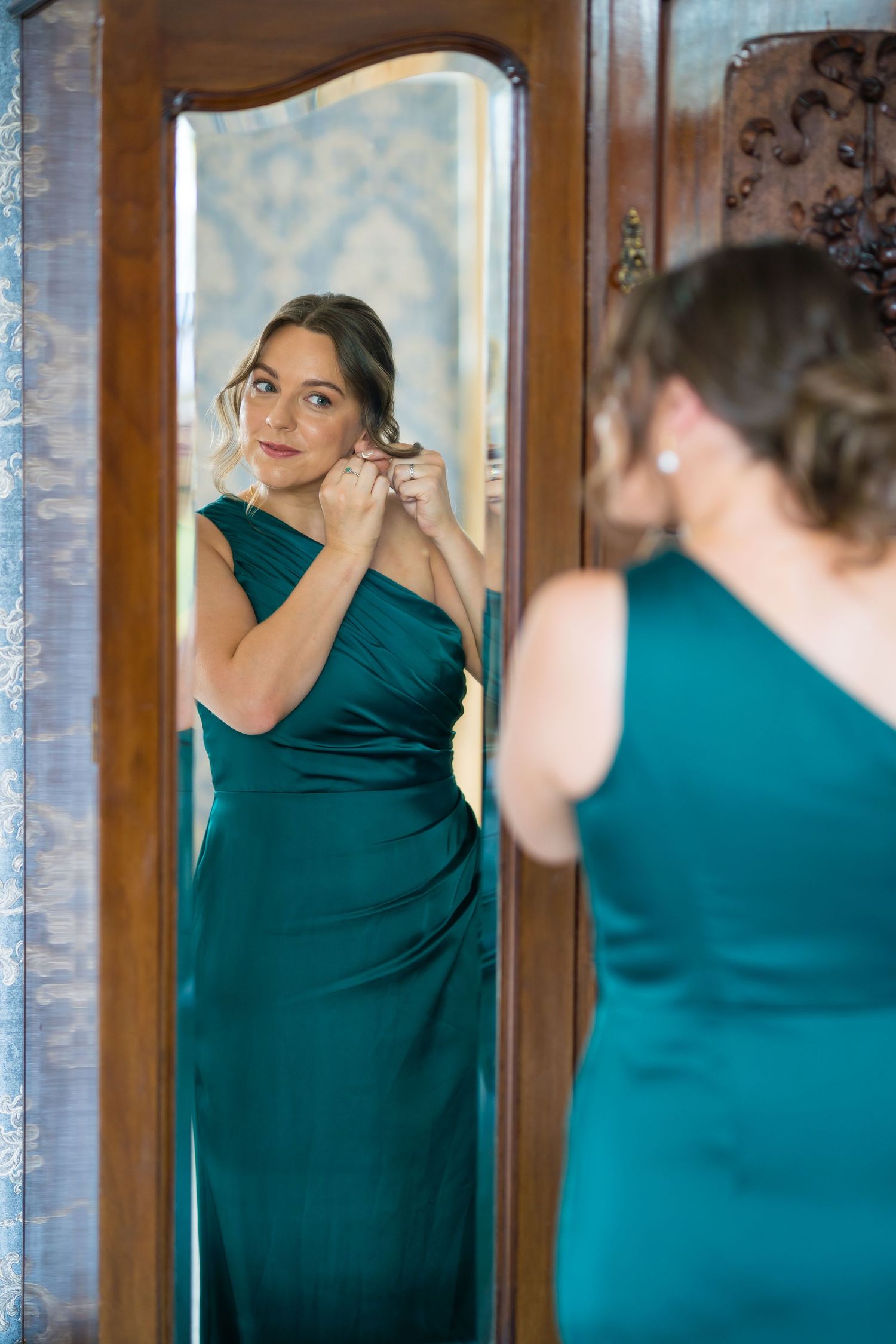Woman in emerald green gown adjusting earring while looking in ornate mirror.