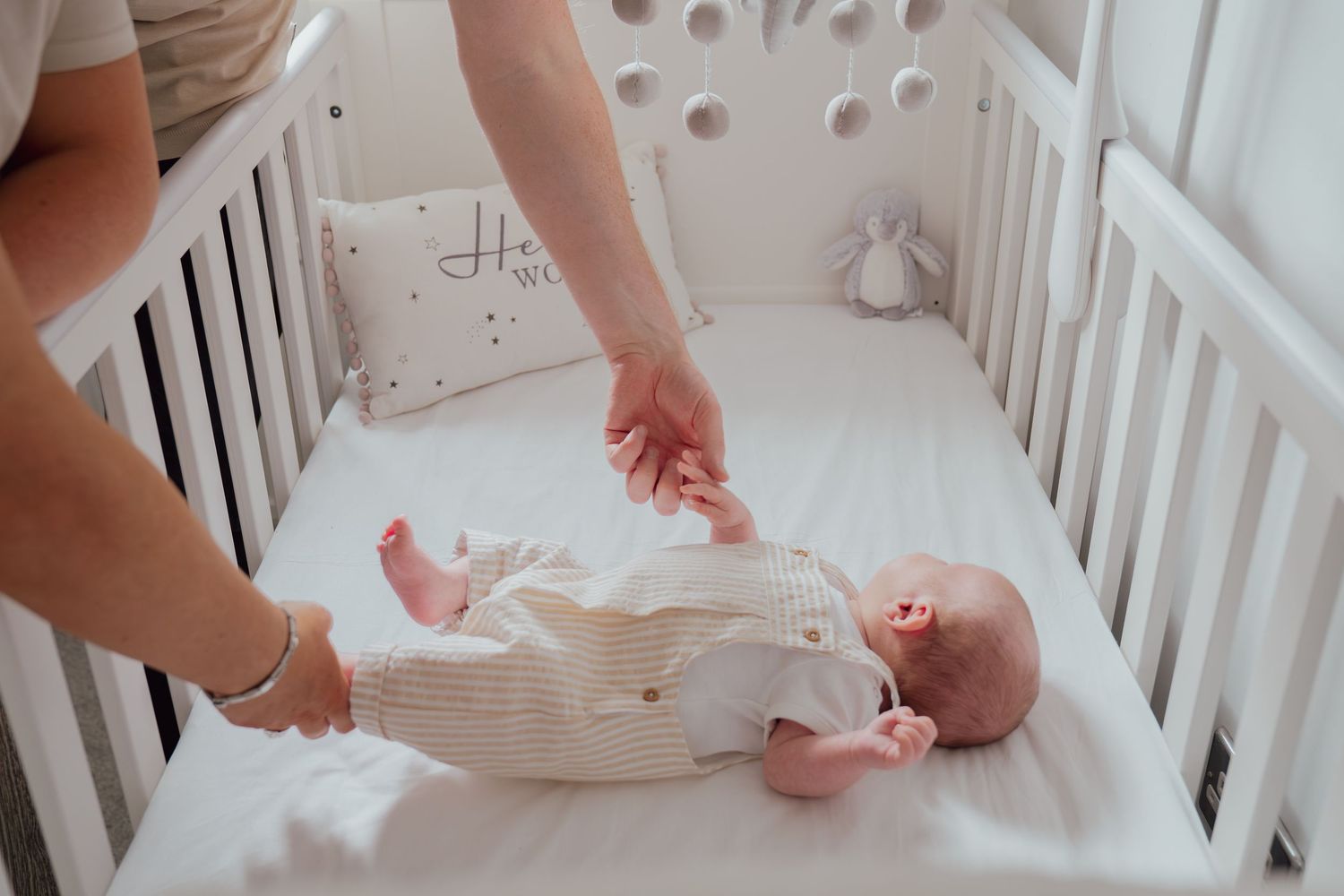 Parent gently holding newborn baby's hand while lying in a white wooden crib with soft bedding.