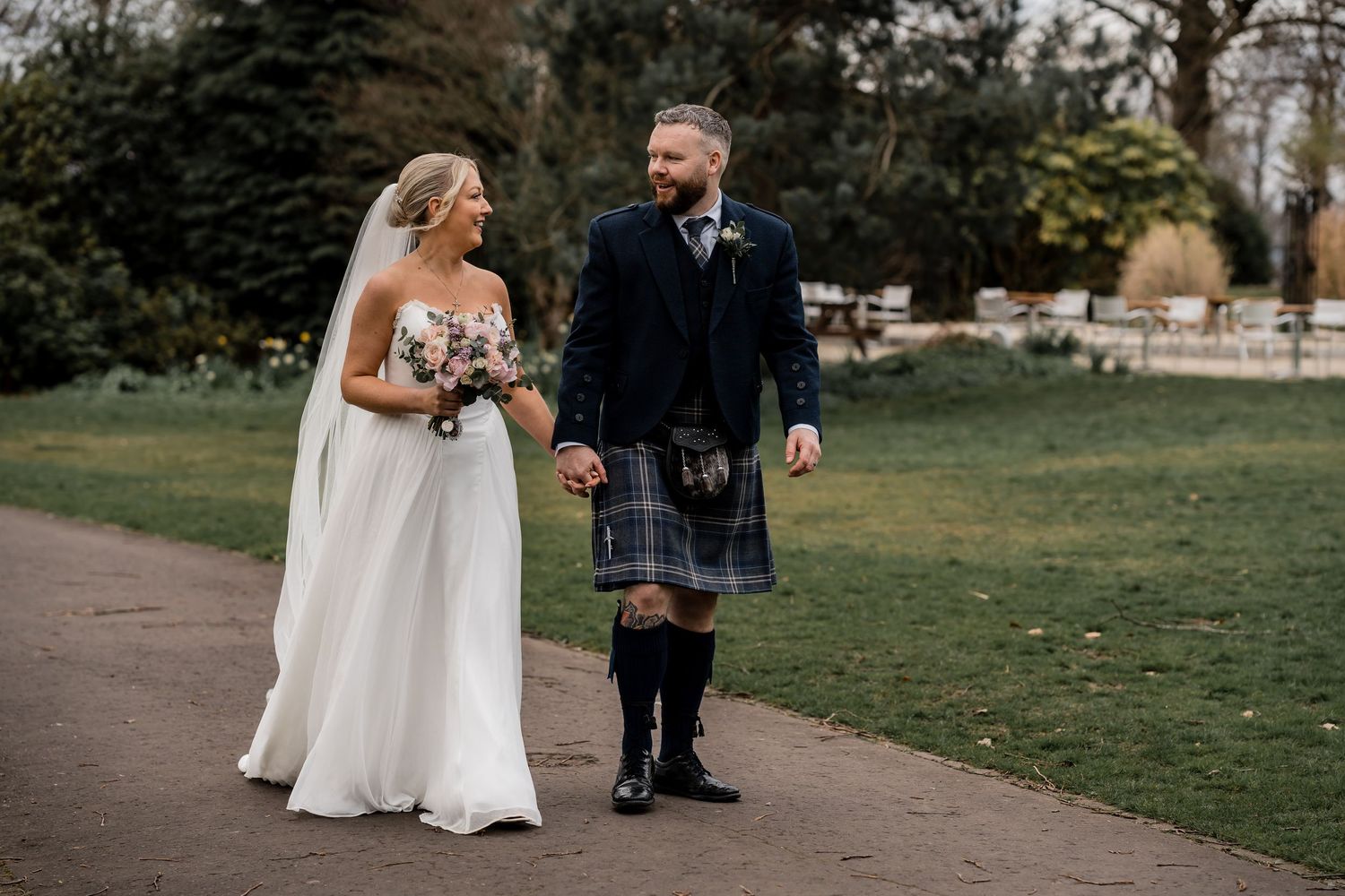 Bride and groom walking hand in hand in a park, groom wearing a traditional Scottish kilt, bride in a white wedding gown.
