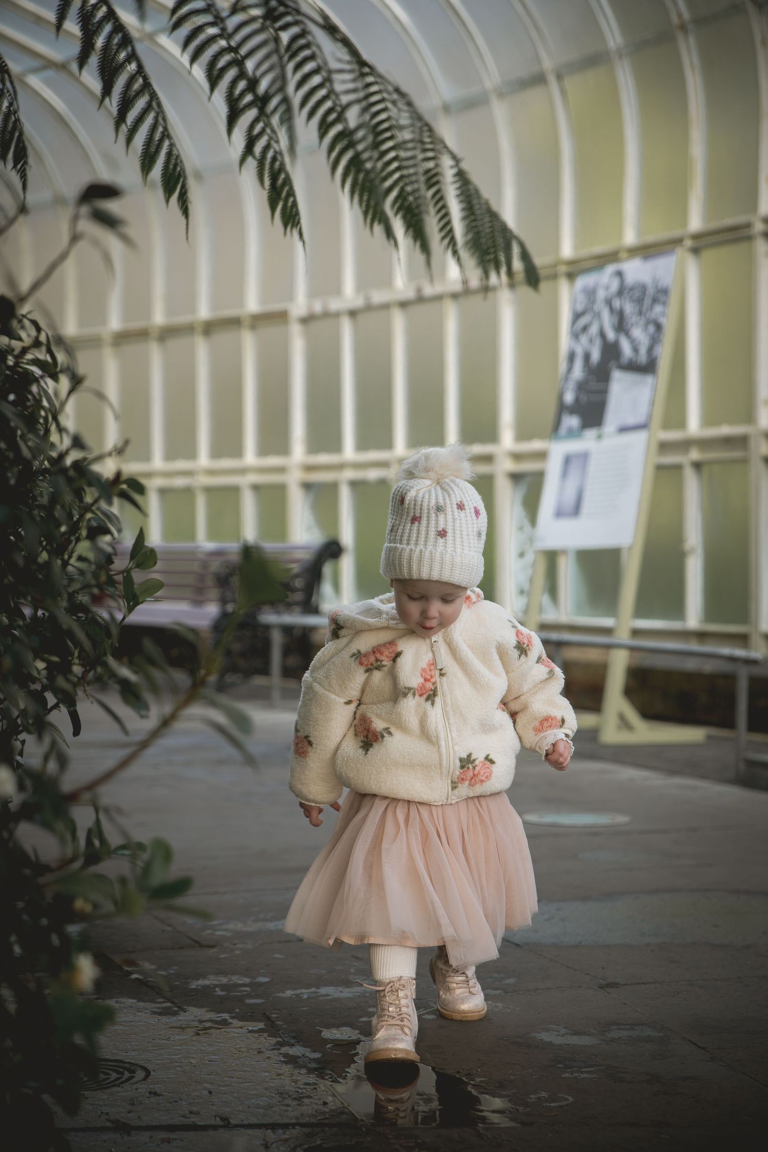 Toddler girl in floral sweater, pink tutu skirt, and white pom-pom hat walking in a greenhouse.