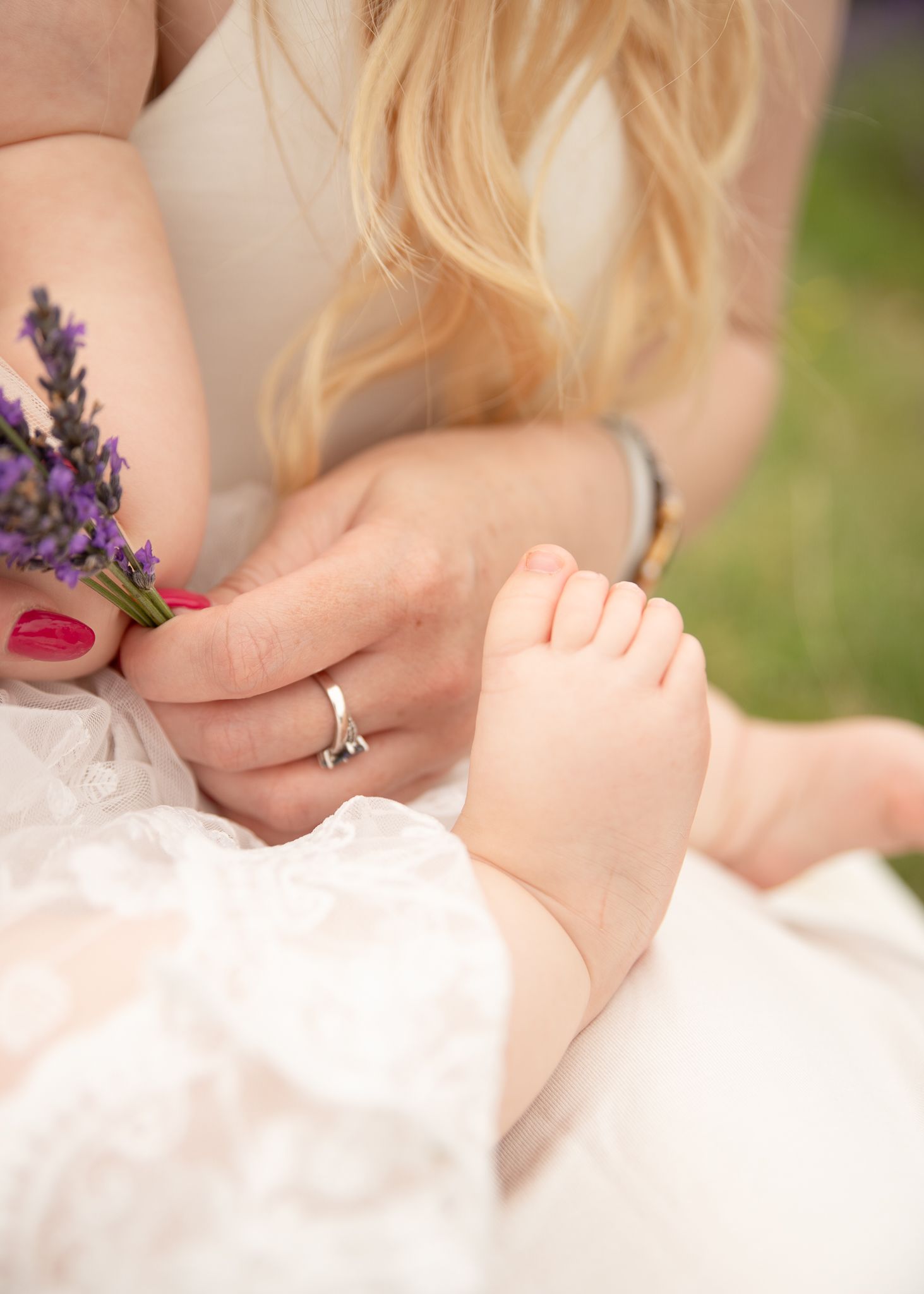 Blonde woman in white lace dress holding lavender flowers with a baby's tiny foot resting on her lap.