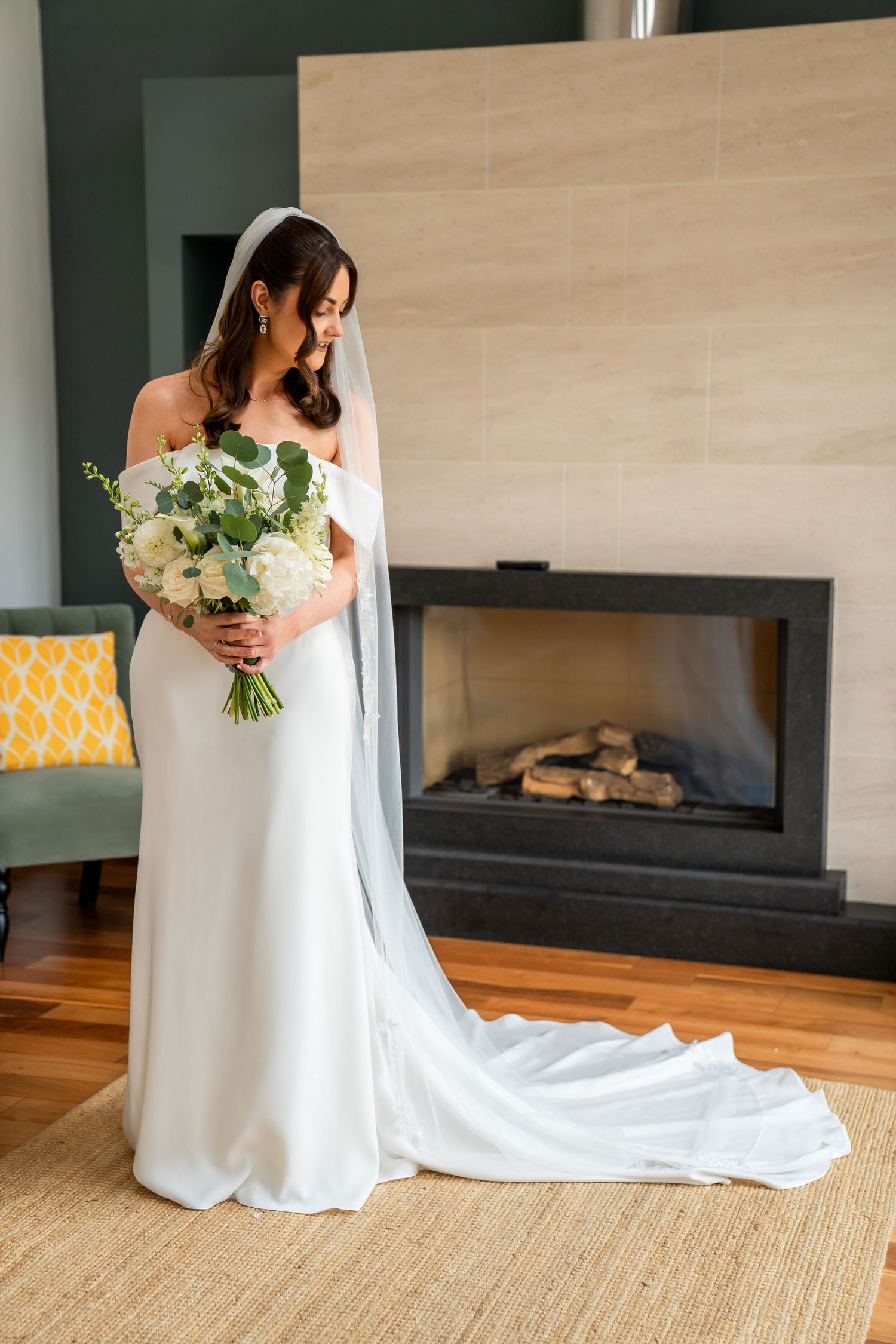 Elegant bride in off-shoulder white gown holding white floral bouquet standing before a modern fireplace.