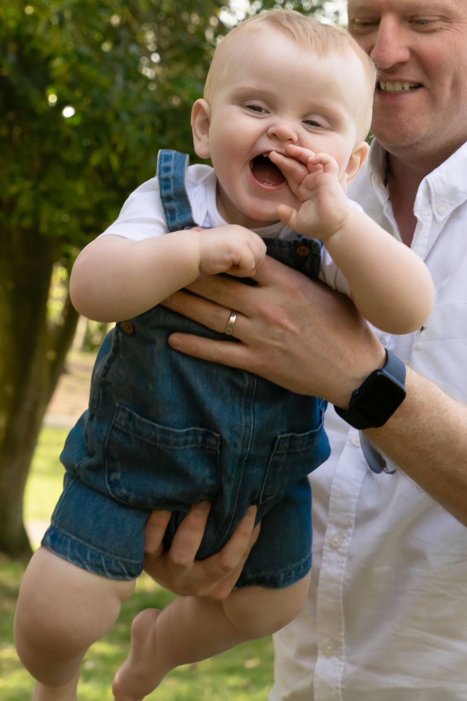 A laughing baby in denim overalls being held by a smiling man in a white shirt outdoors in a park.