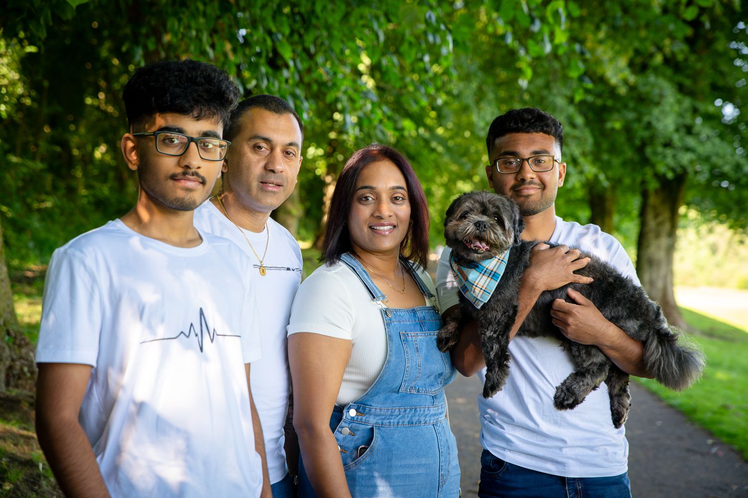 Family of four posing outdoors on a park path, with one member holding a small dark fluffy dog, surrounded by lush green trees.