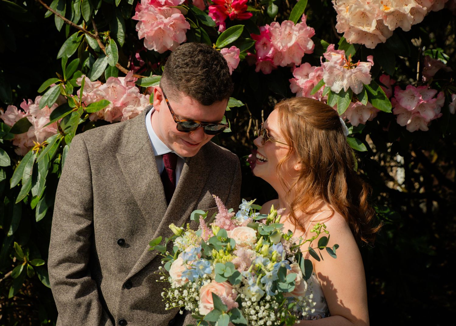 A romantic couple poses among blooming pink roses with a beautiful bouquet.