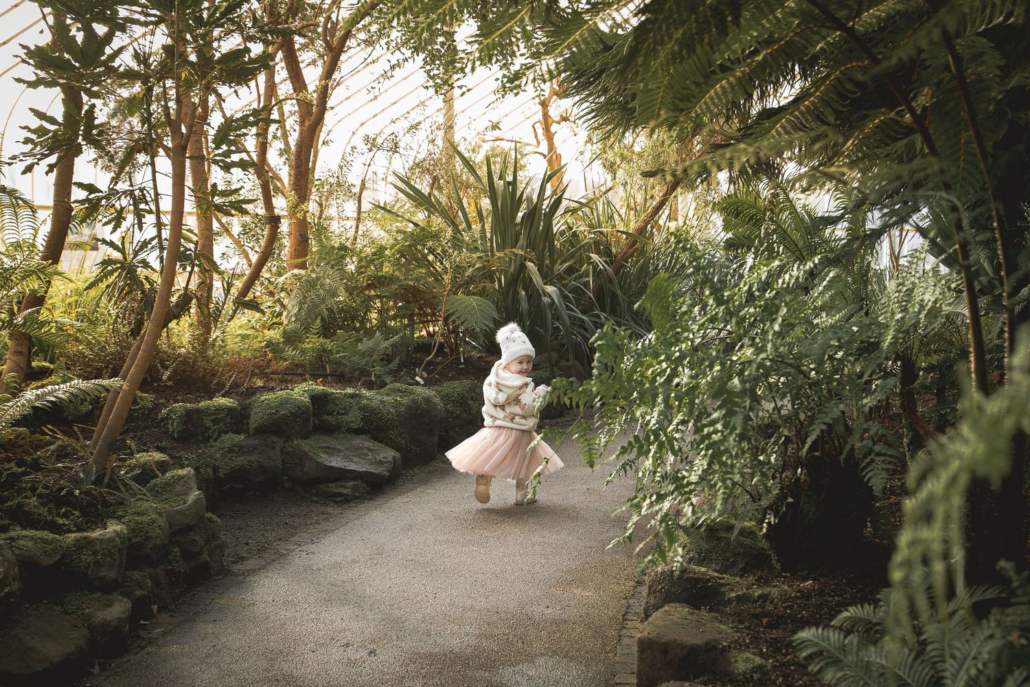 A child in a pink skirt walks along a path surrounded by lush tropical plants inside a greenhouse.