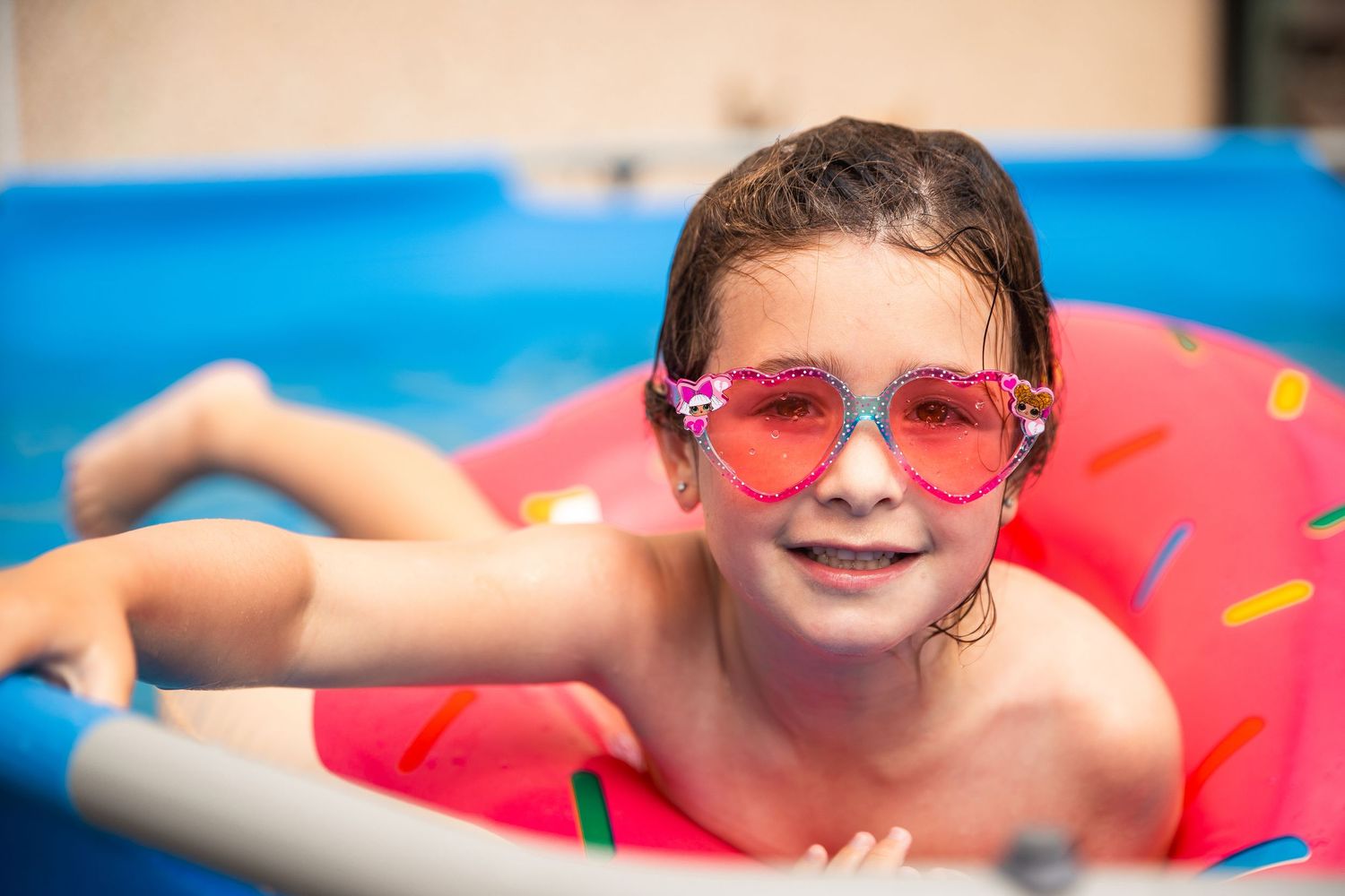 Young girl wearing pink heart sunglasses floating on a donut inflatable ring in a backyard swimming pool.