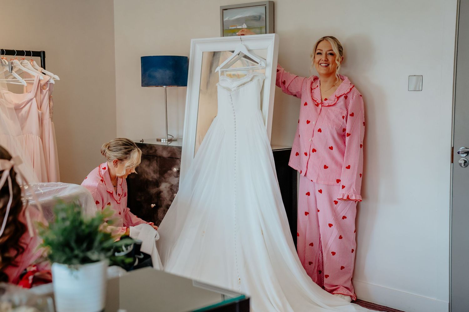 Two women in pink pajamas prepare a white wedding dress hanging in a bright room on wedding morning.