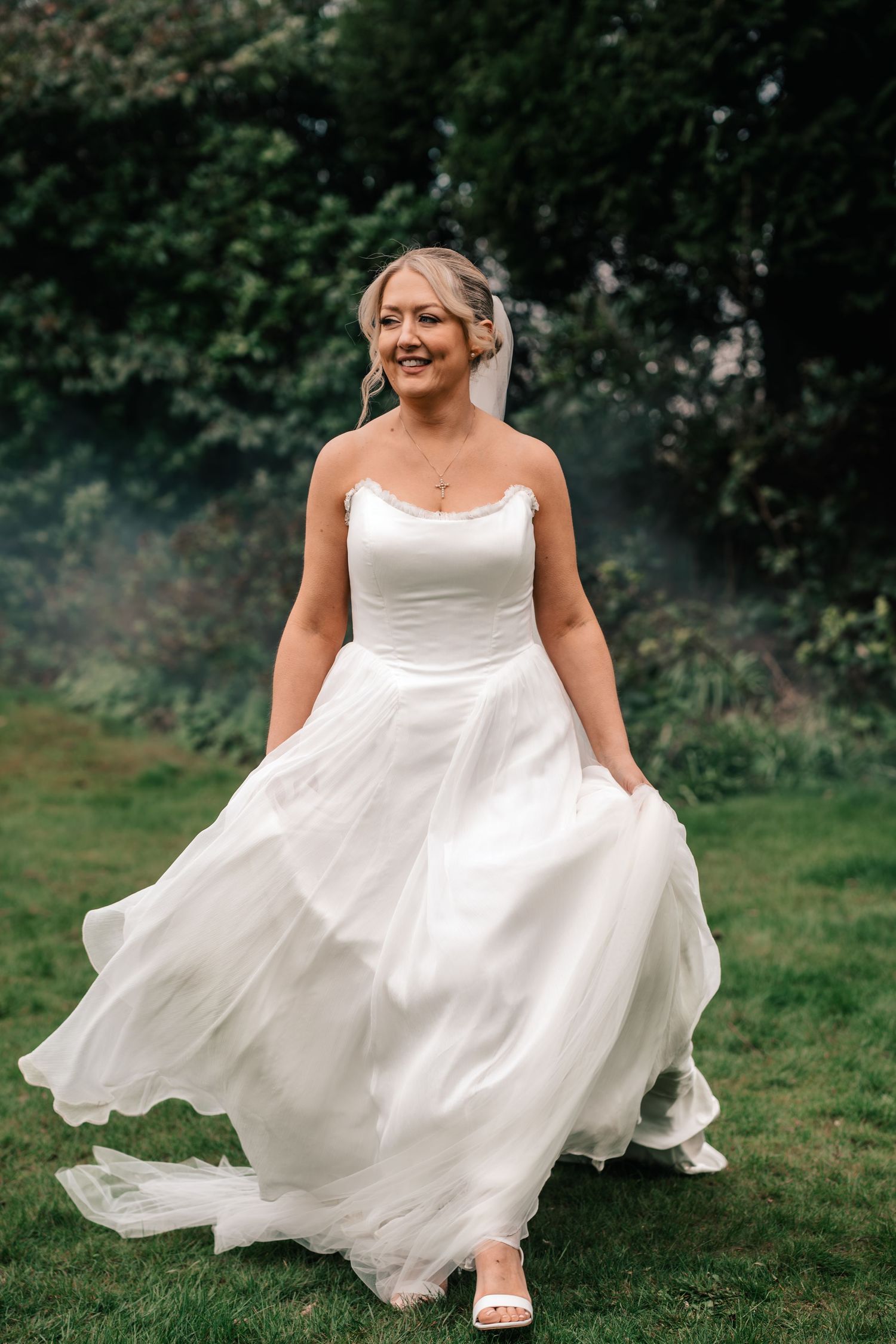 Smiling bride in a flowing white ball gown walks outdoors on green grass with trees in the background.