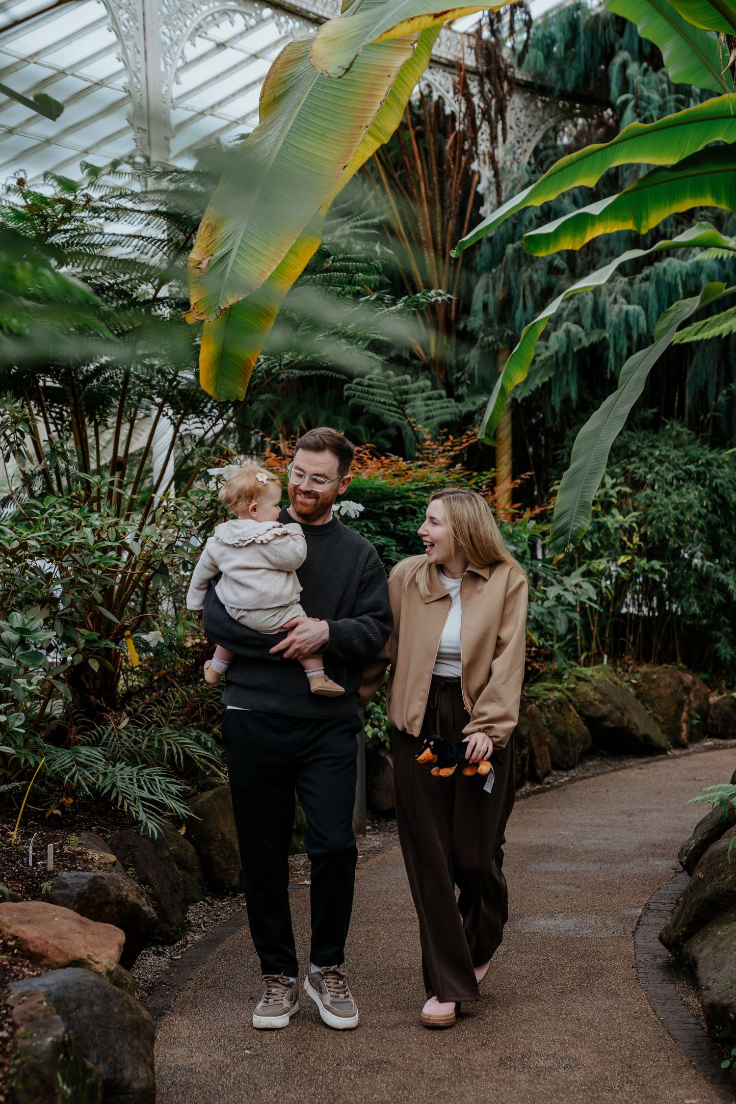 A young family with a baby strolls through a lush tropical greenhouse, smiling warmly among large green plants.