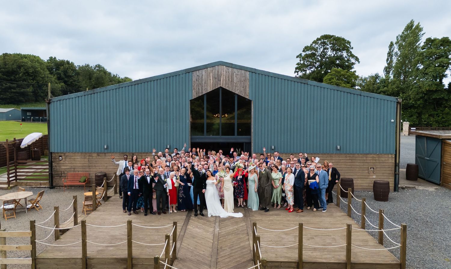 Large wedding group photo outside a rustic barn venue with wooden deck, surrounded by lush greenery.