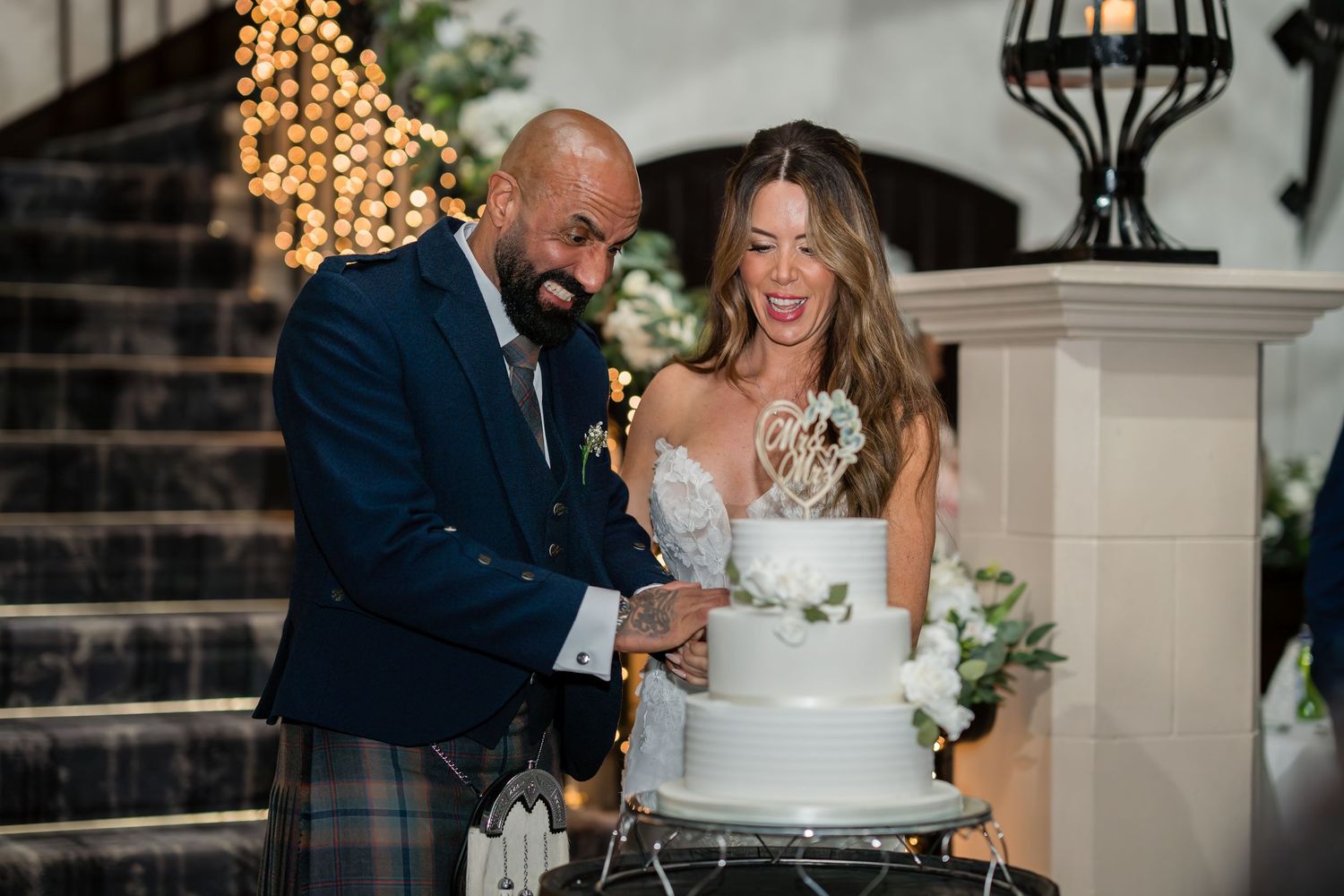 A couple cuts their elegant white wedding cake together while laughing during their reception celebration.