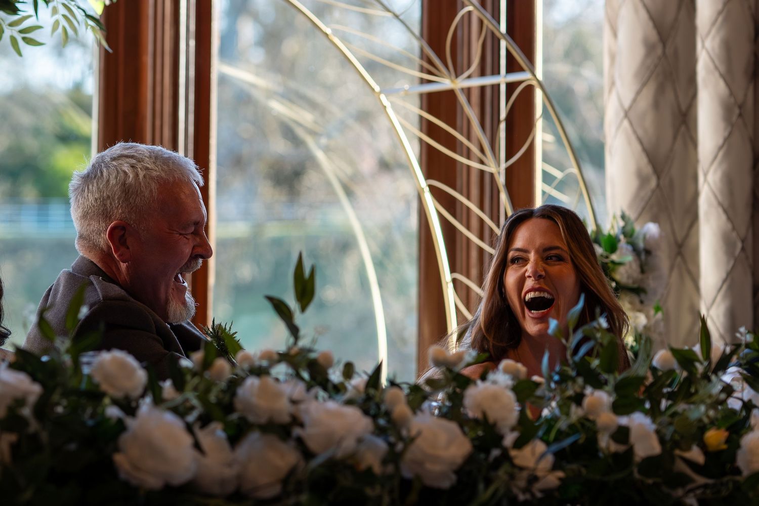 A joyful moment captured during a wedding reception with white floral decorations and natural window lighting.