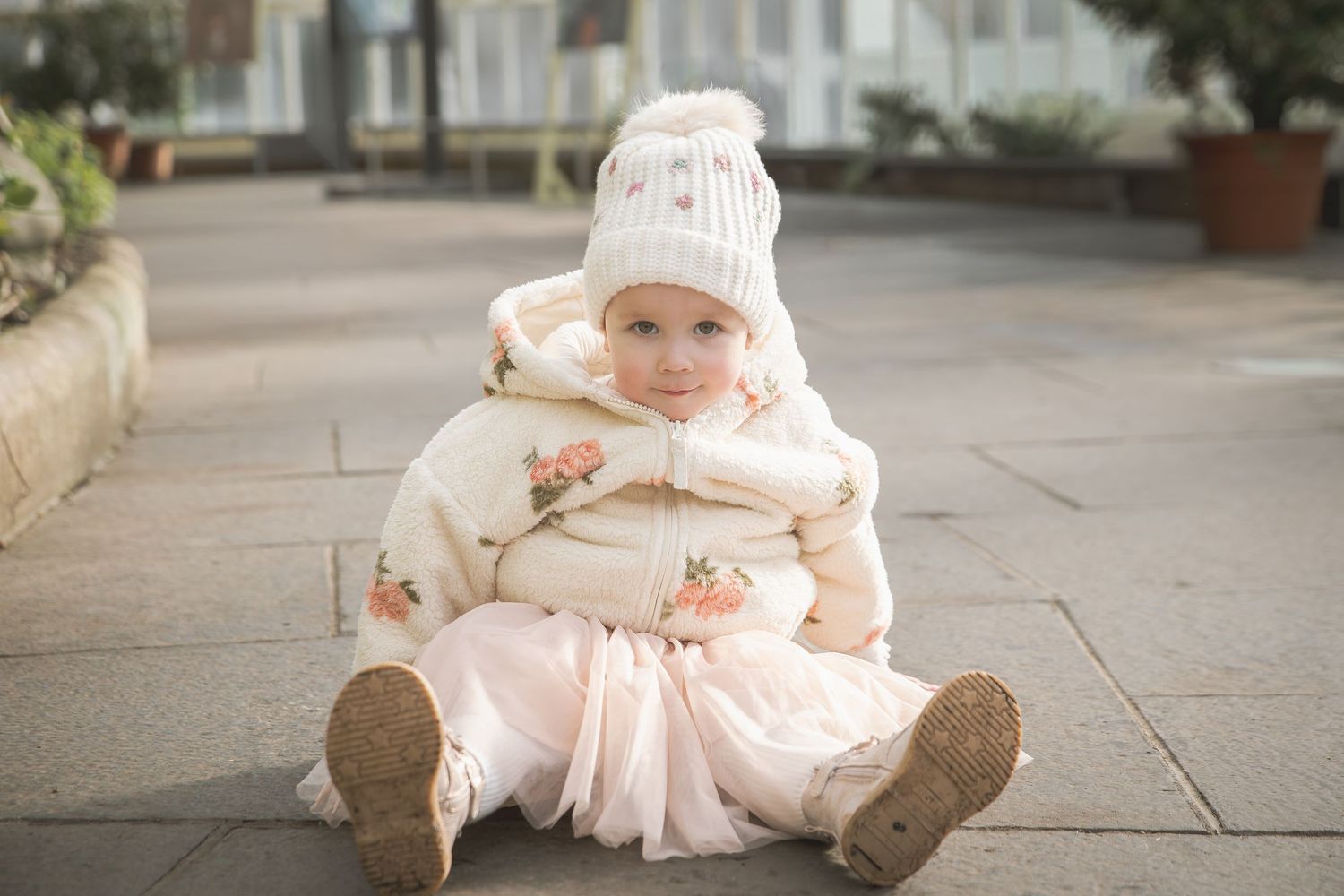 Adorable toddler girl in white floral winter jacket, pink tutu skirt, and knit hat sitting on pavement outdoors.