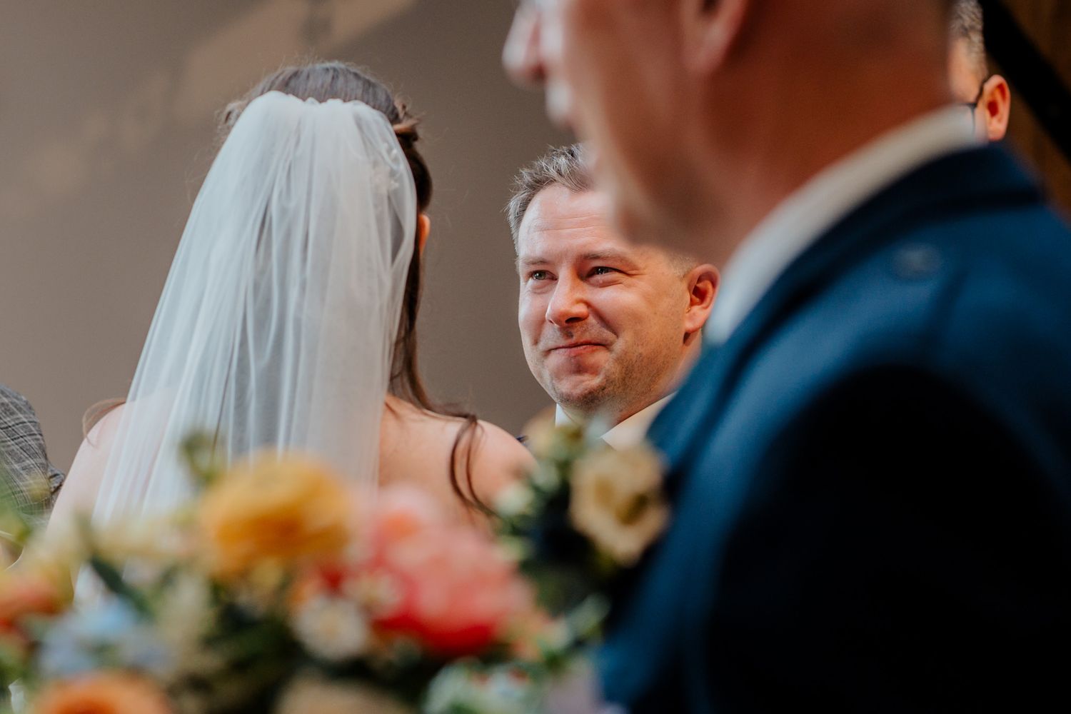 Groom smiling emotionally at bride during wedding ceremony with colorful floral arrangements in foreground.