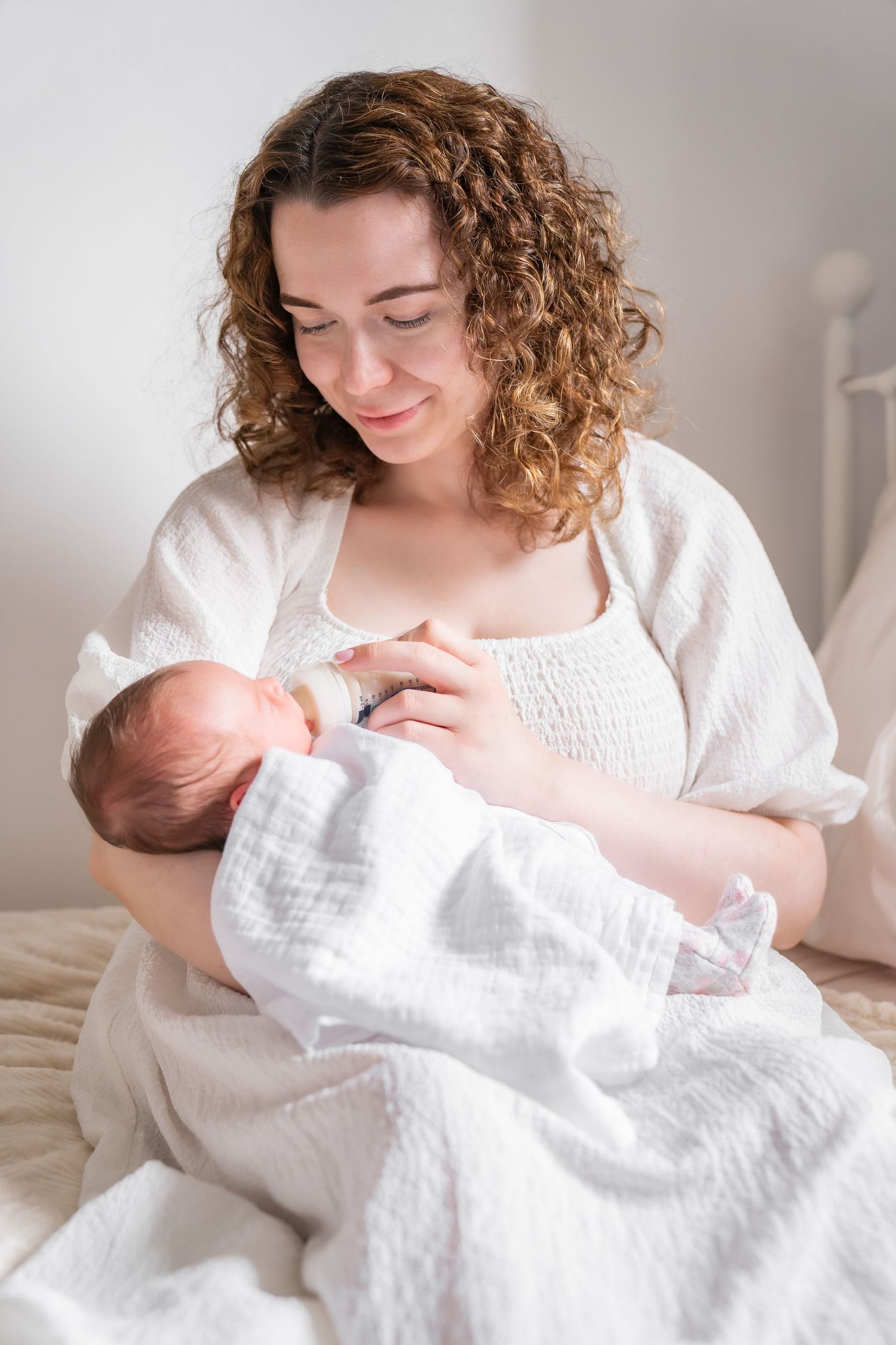 A smiling mother with curly hair bottle-feeding her newborn baby wrapped in a white blanket while sitting on a bed.