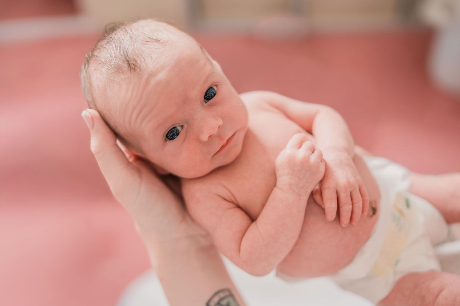 Newborn baby being held in a tattooed hand, alert with blue eyes, wrapped in a white blanket in a hospital.