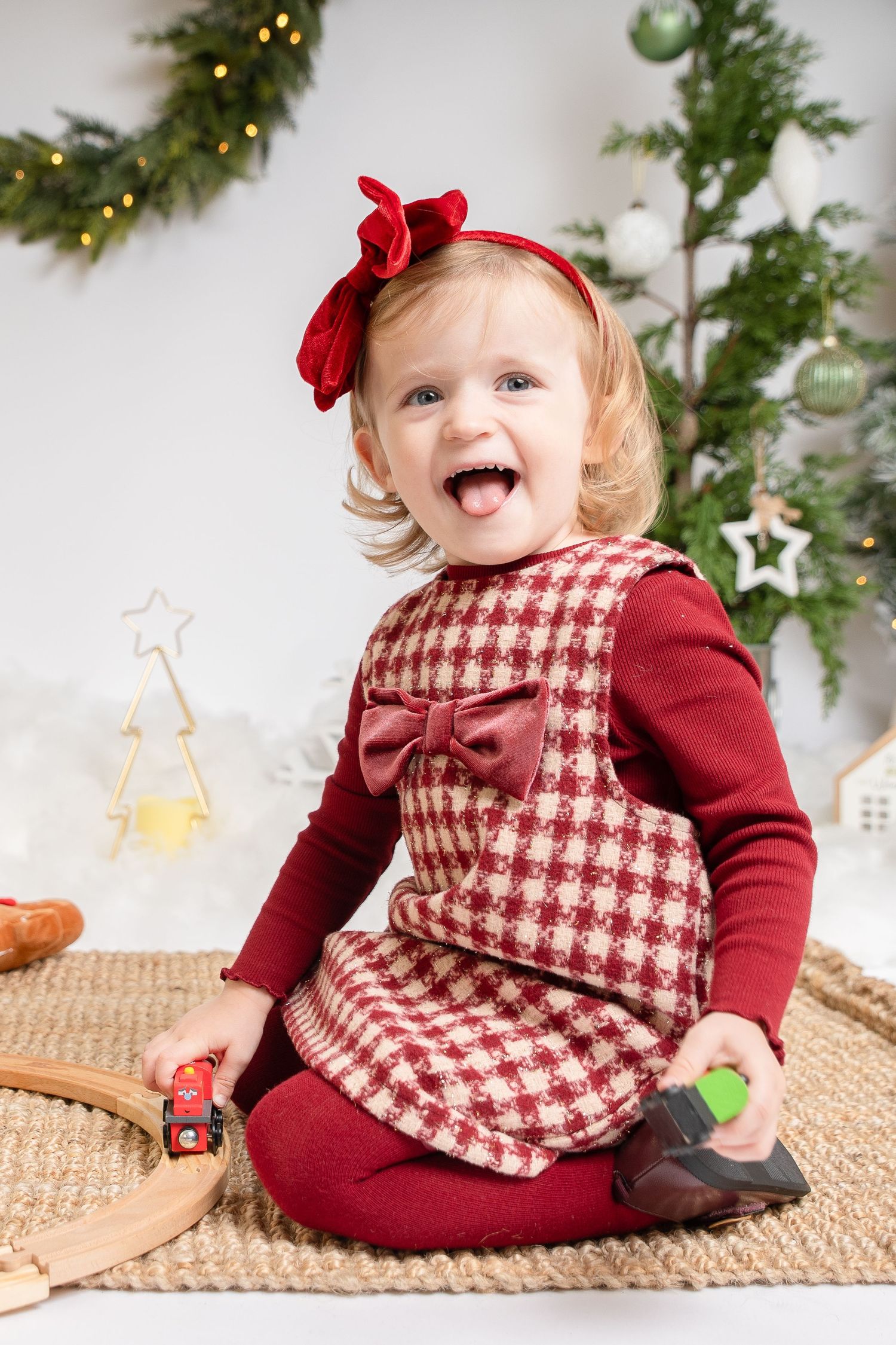 Cheerful toddler girl in red plaid Christmas dress with bow headband sitting by decorated tree with toys.