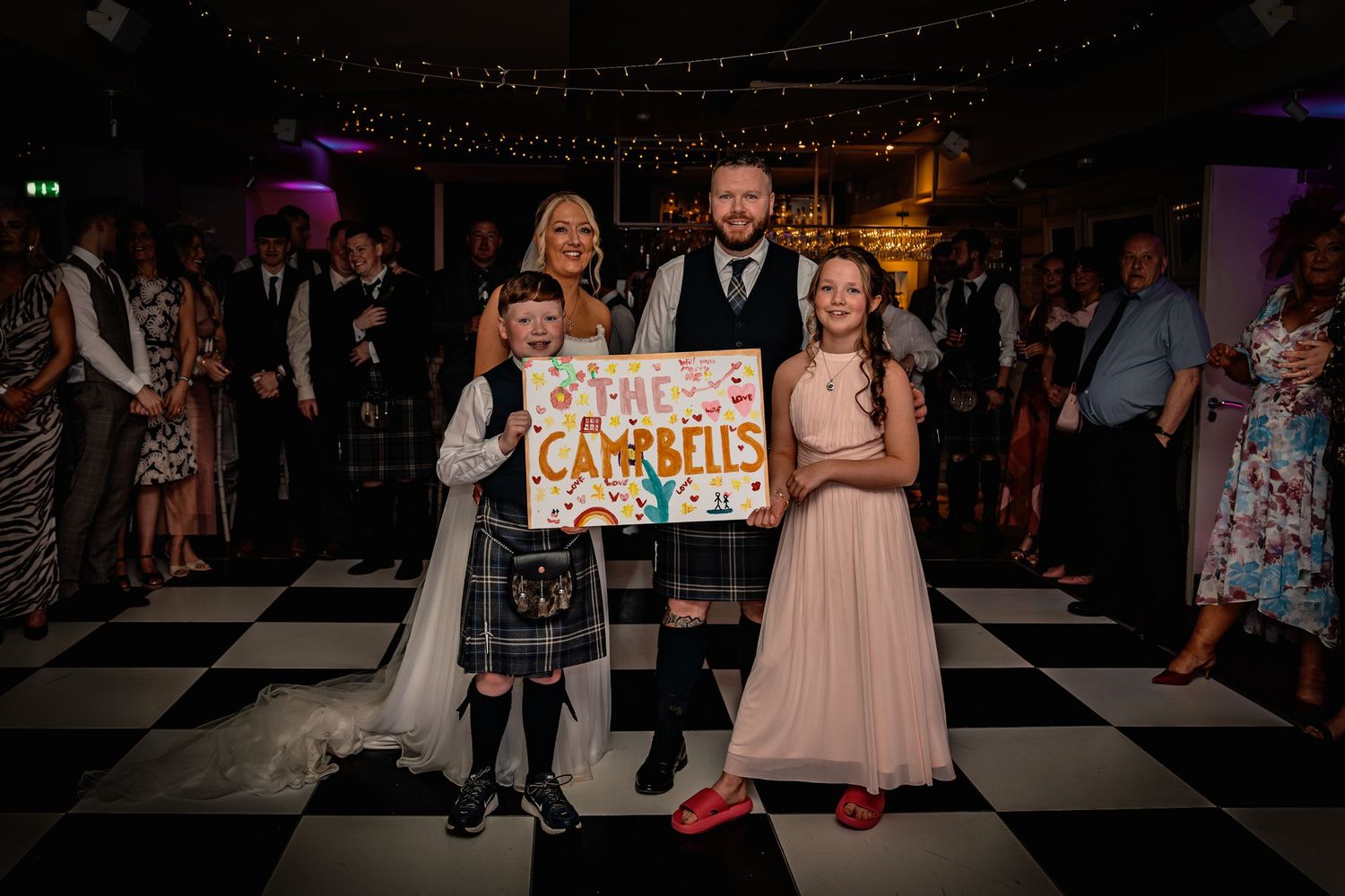 Family posing with The Campbells wedding sign on black and white dance floor at Scottish wedding reception.
