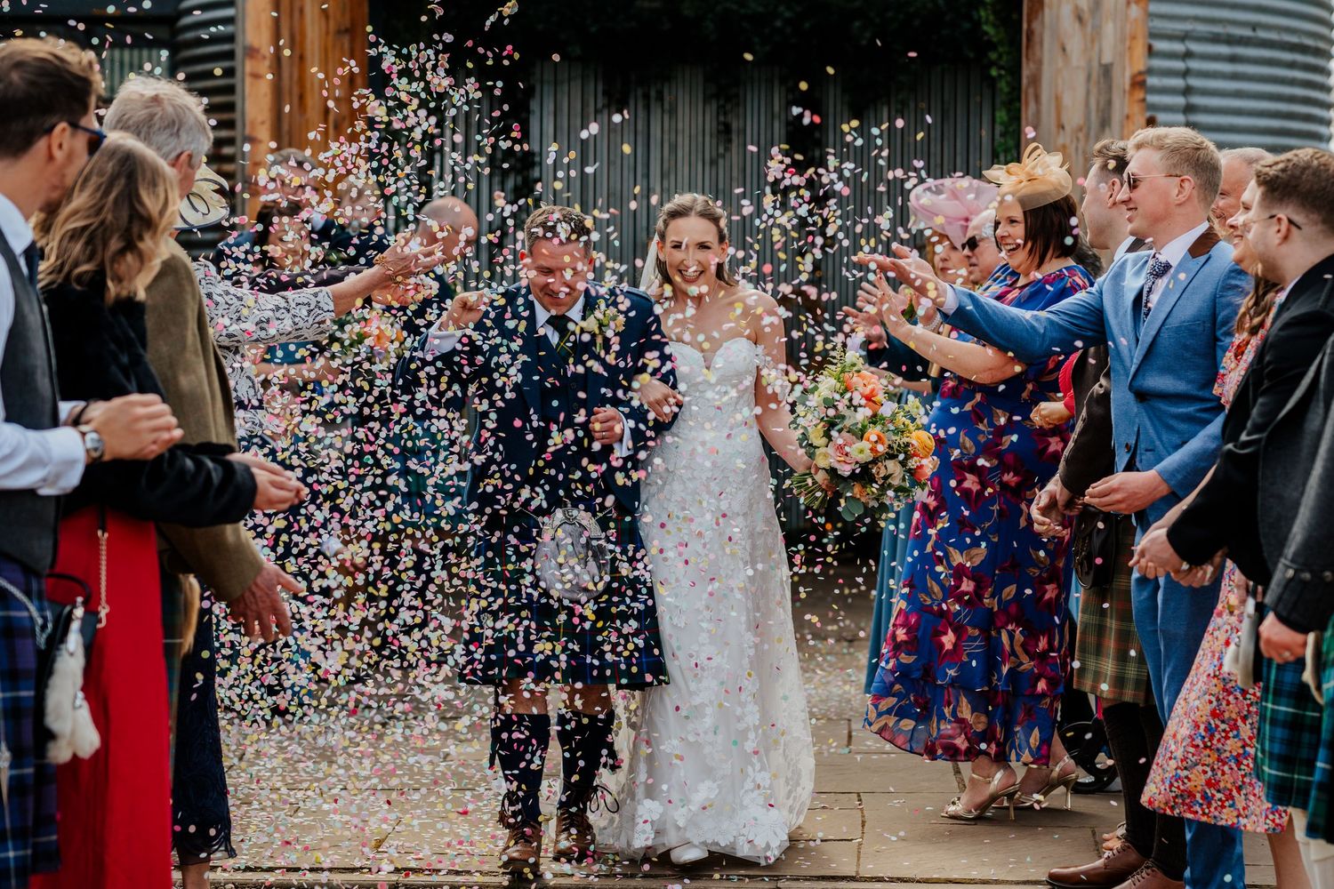 Bride and groom walk through colorful confetti shower surrounded by cheering wedding guests outside a rustic barn venue.