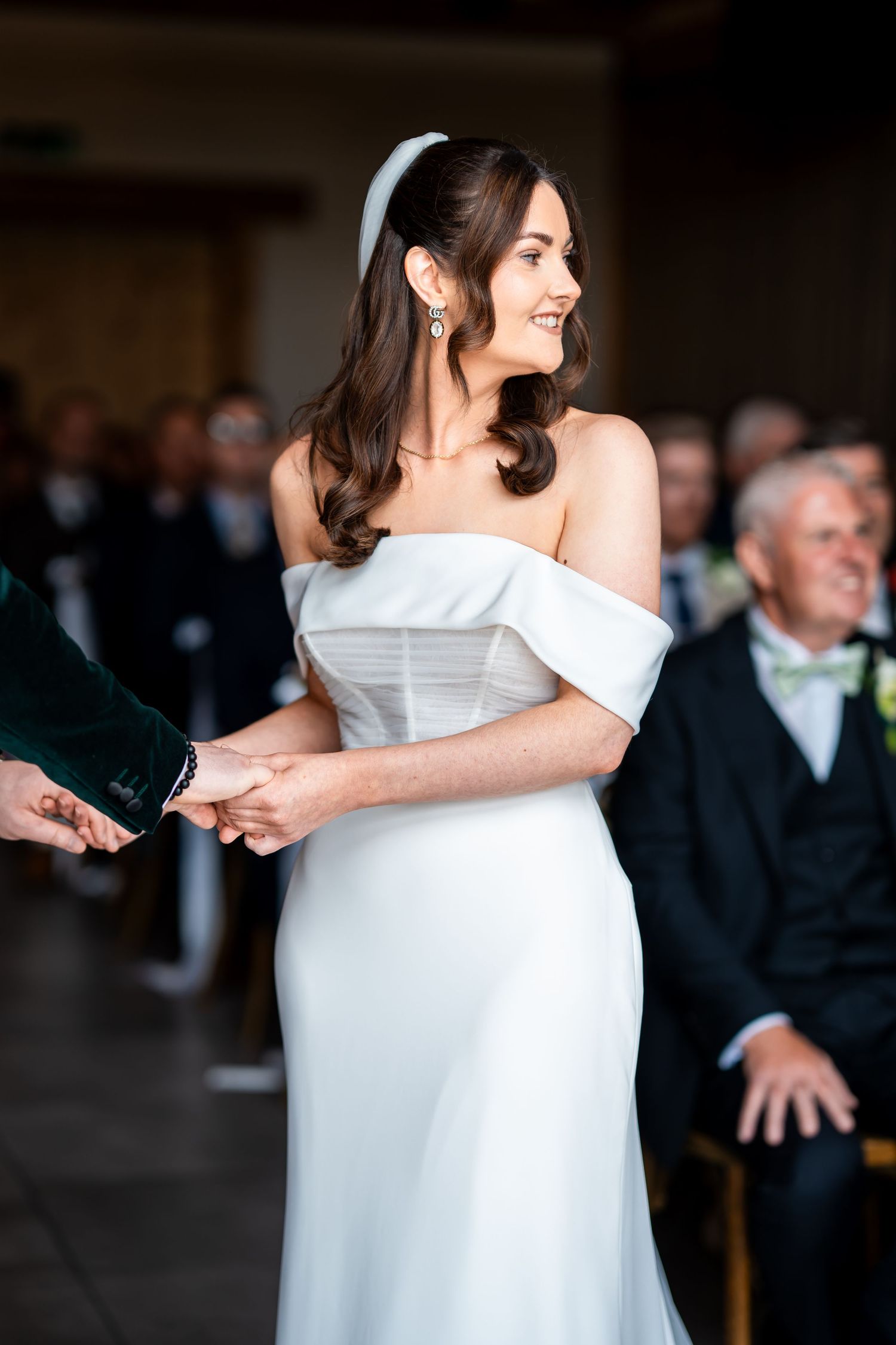 Bride in off-shoulder white gown holding hands at wedding ceremony, smiling joyfully with guests seated behind her.