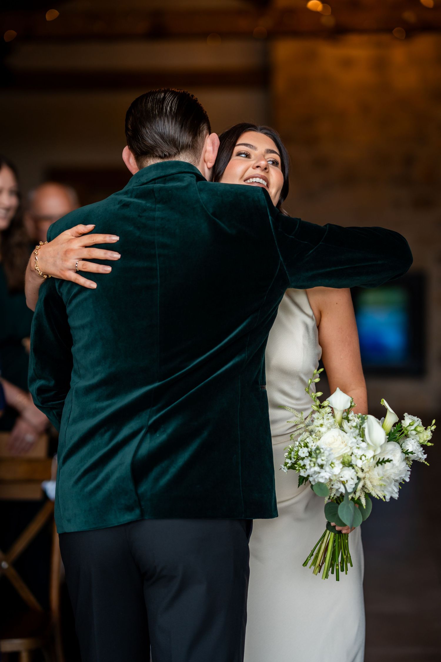 Bride in white gown hugging groom in green velvet blazer while holding white floral bouquet during wedding ceremony.