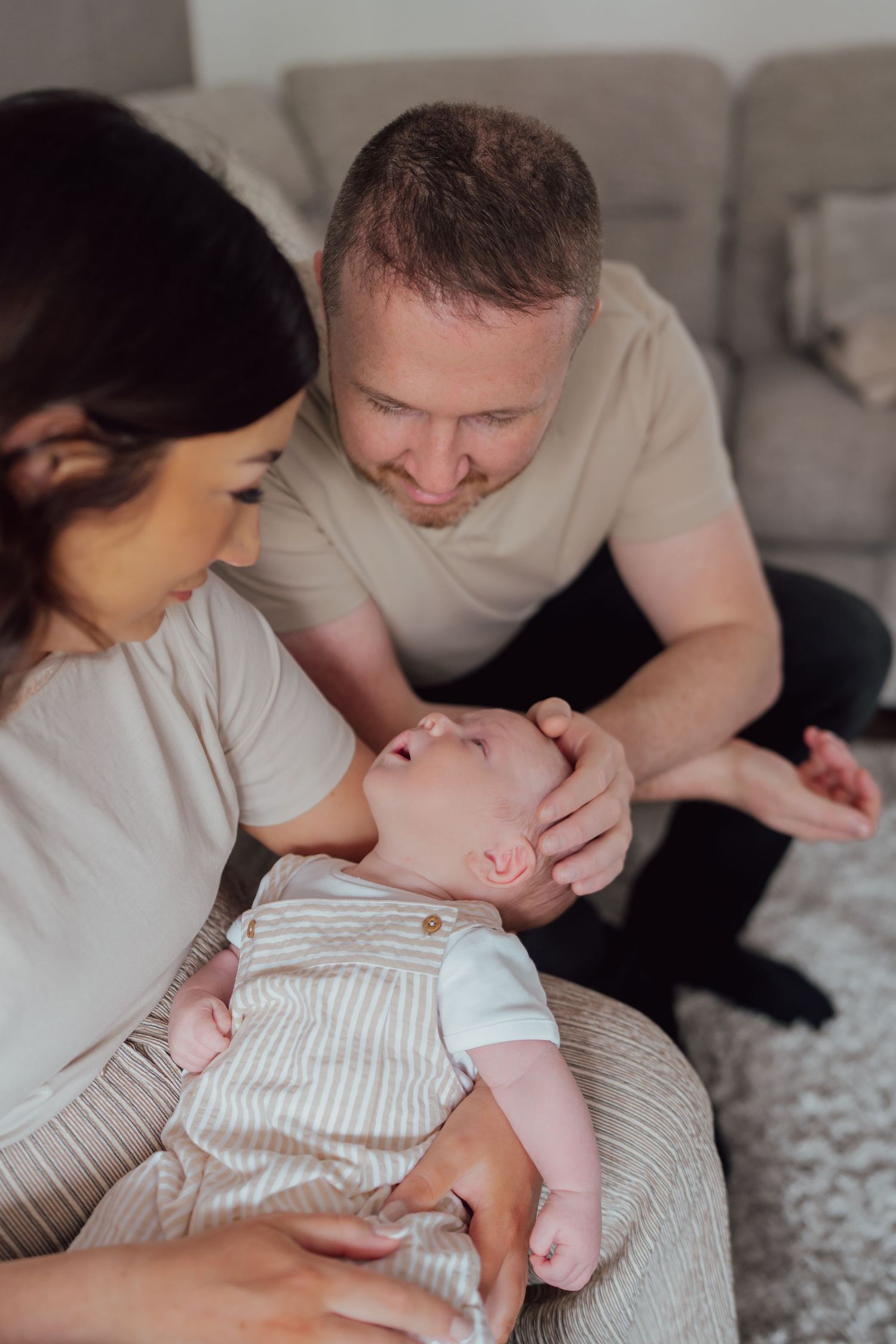 A couple smiles lovingly at their newborn baby while sitting together on a couch in a cozy living room.