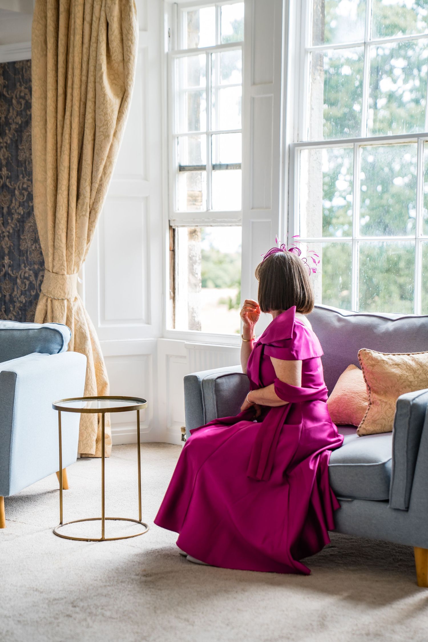 Woman in magenta gown and floral fascinator sits on grey sofa gazing out large window in elegant room.