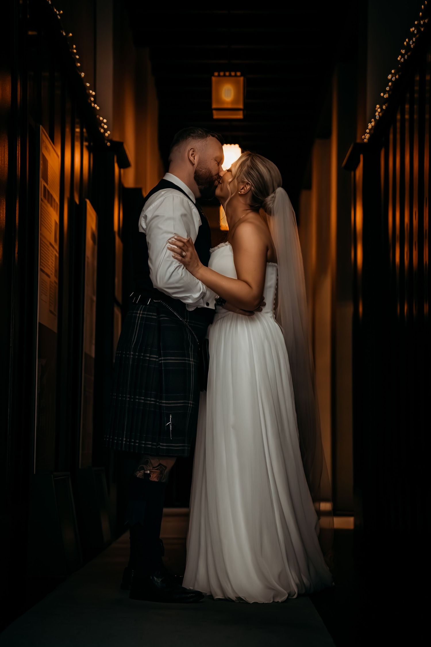 Bride and groom sharing a romantic kiss in a dimly lit corridor, groom wearing a traditional Scottish kilt.
