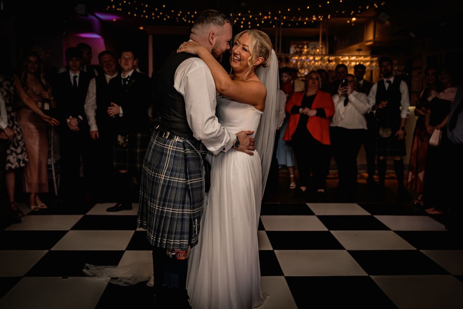 Bride and groom share first dance on checkered floor, groom in Scottish kilt, guests watching in warmly lit wedding venue.