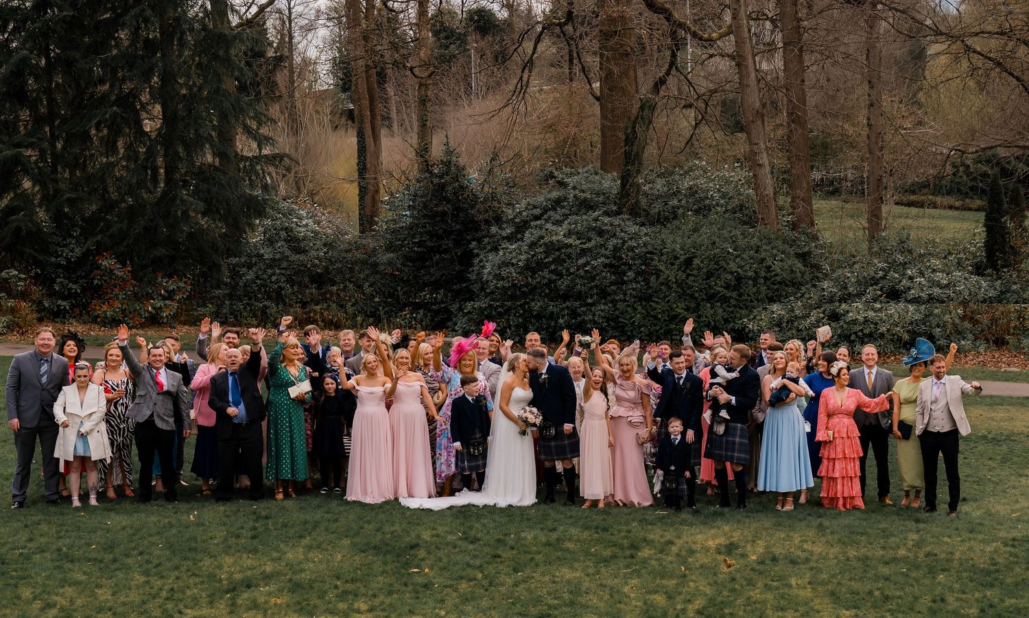 Large wedding party group photo outdoors on green lawn with trees, bride in white gown surrounded by guests in colorful attire.