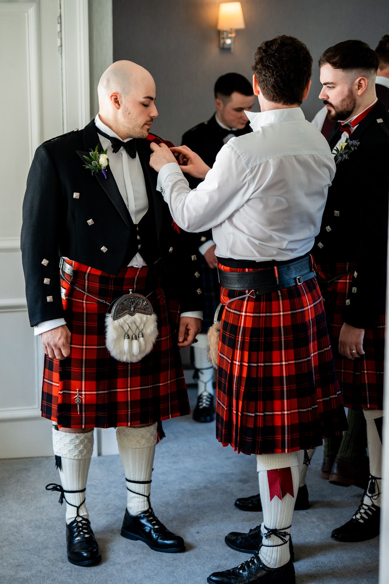 Men in traditional Scottish kilts with red tartan prepare for a wedding ceremony indoors.