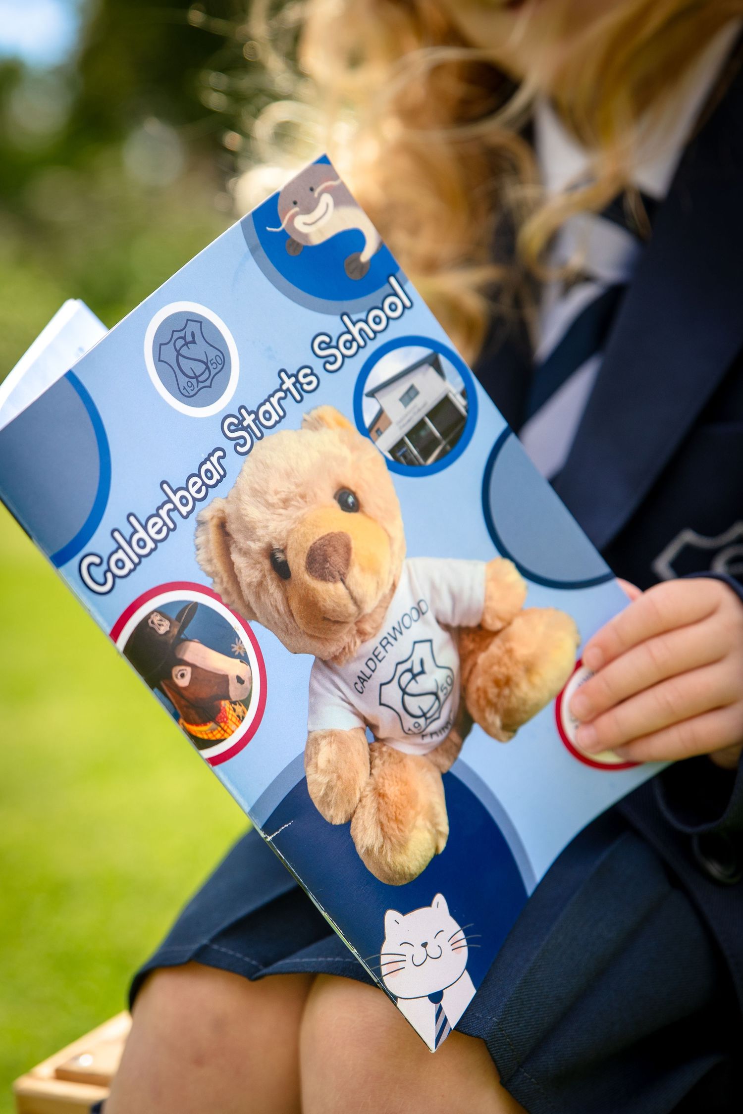 A child in school uniform holds a book titled Calderbear Starts School featuring a teddy bear mascot.