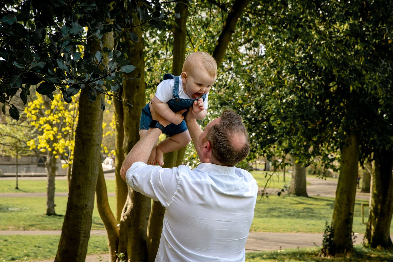A man lifts a happy baby boy into the air in a lush green park surrounded by trees.