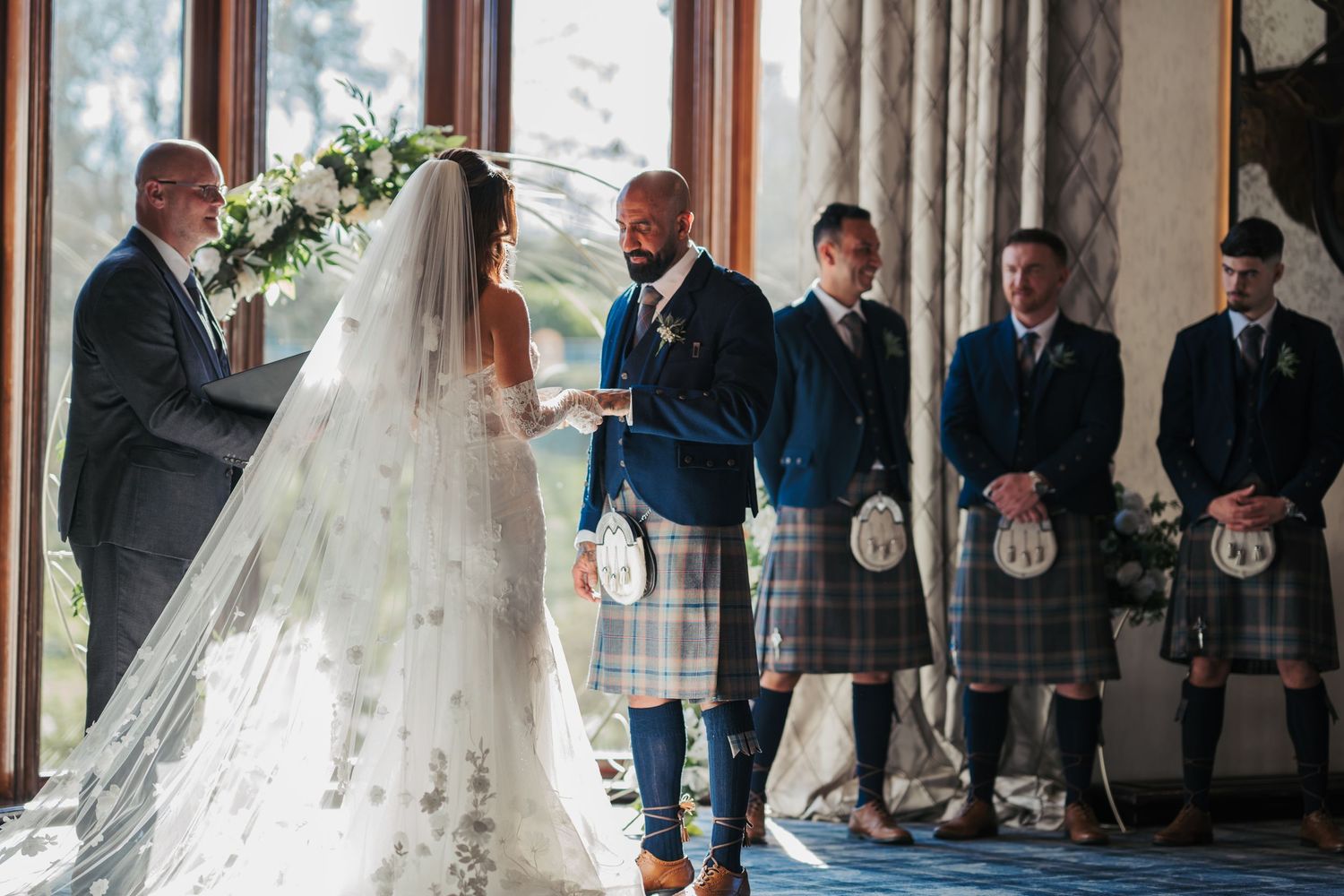 A bride and groom stand in front of their wedding party wearing traditional Scottish kilts during a formal ceremony.