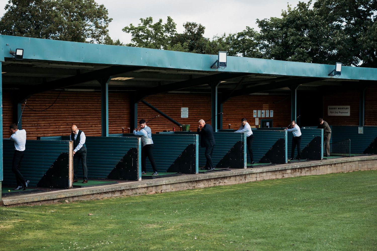 People standing along a teal-painted covered grandstand at a sports ground, surrounded by green grass and trees.